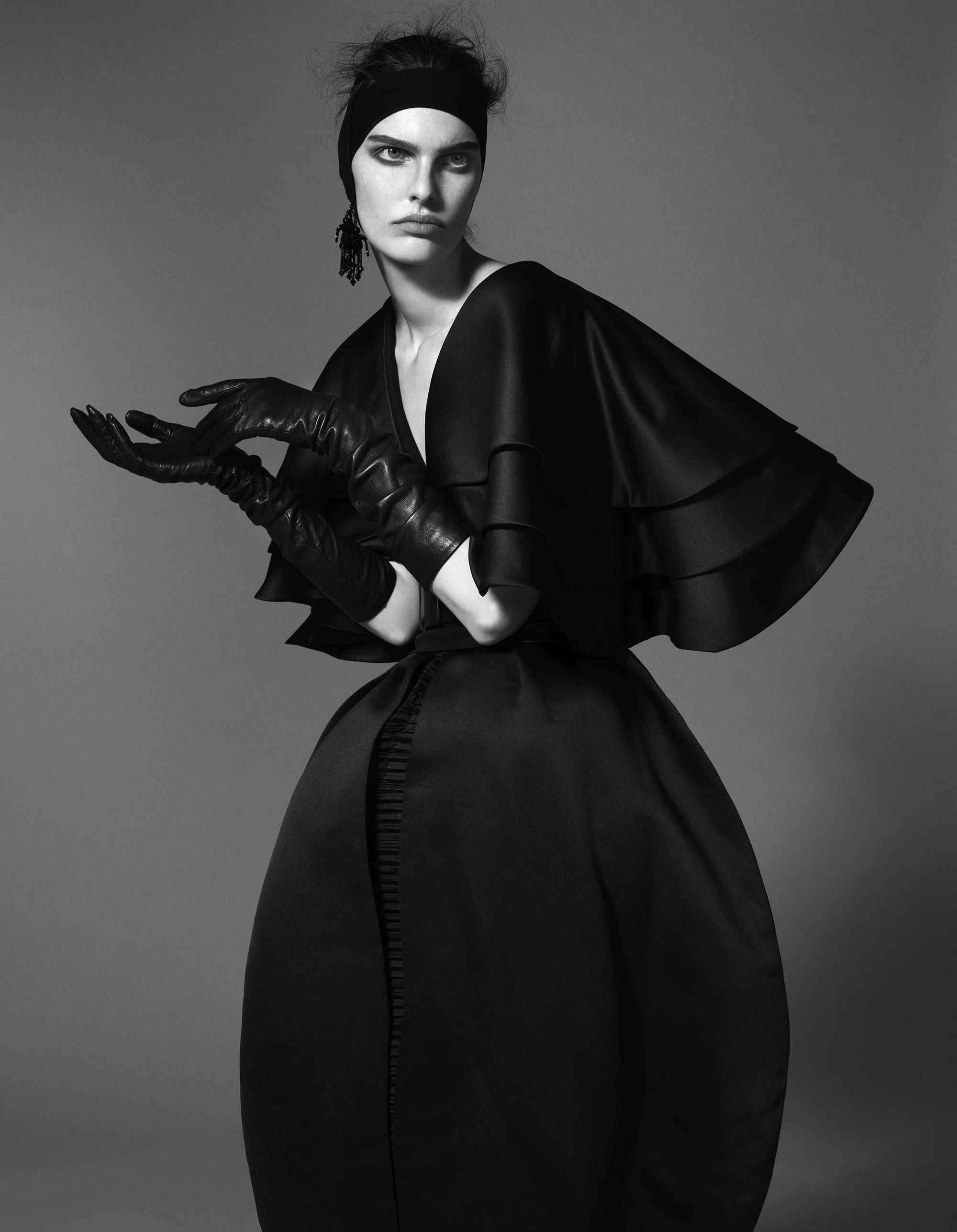 Black and white photograph of a woman in a dramatic pose, wearing black leather gloves, a glossy black dress with ruffled sleeves, a black headband, and a statement earring. Her gaze is intense and her hair is styled in an updo. The background is plain and gradient from dark to light, emphasizing her figure.