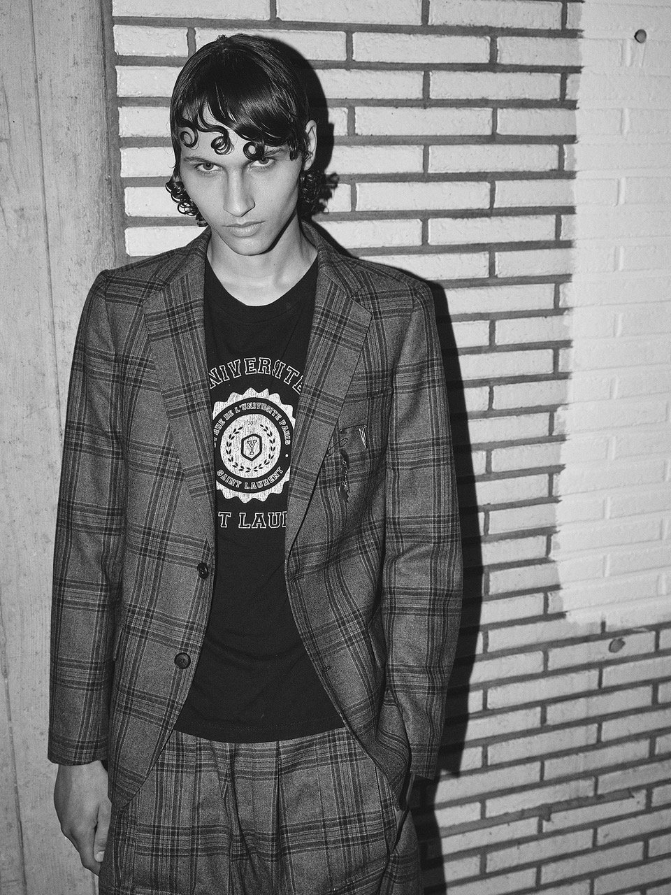 Black and white photo of a young man with curly hair, wearing a plaid blazer and matching plaid pants, standing against a brick wall.
