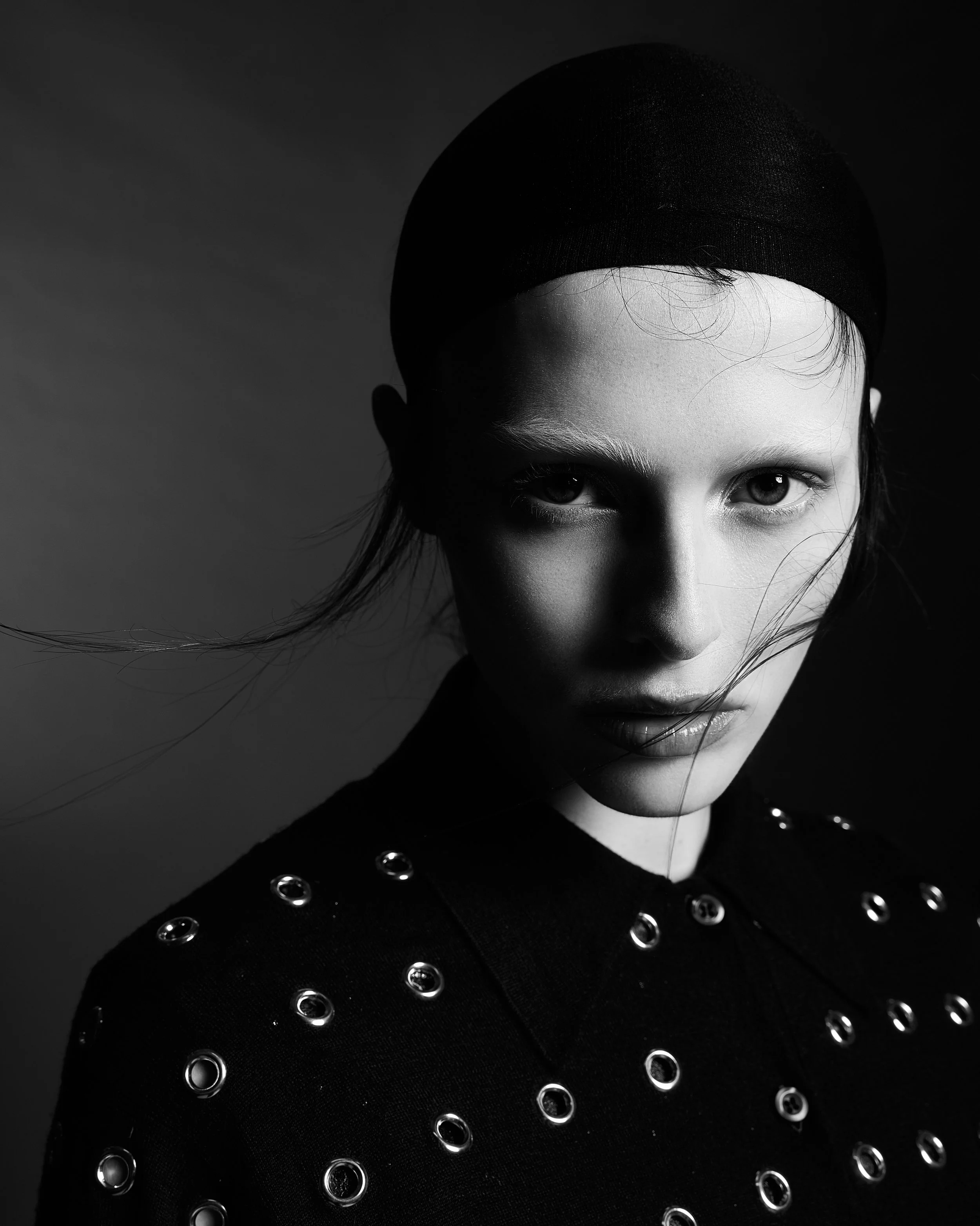 A black and white photograph of a woman wearing a black headscarf and a black top with metallic eyelets, looking directly at the camera.