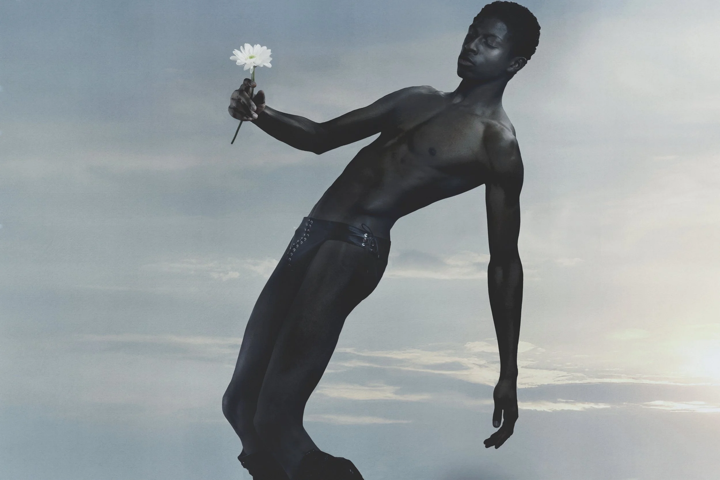 A black person with short curly hair stands shirtless against a gray sky, holding a white flower in one hand while looking at it thoughtfully.