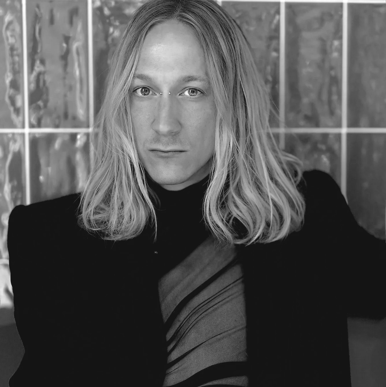 A black-and-white photo of a woman with long, wavy blonde hair, wearing a dark turtleneck sweater, looking directly at the camera with an intense expression, seated in front of a tiled wall.