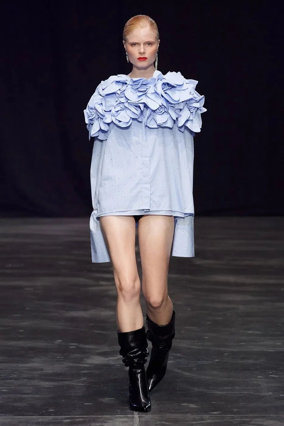 Fashion runway model wearing a light blue shirt with ruffled detailing on the shoulders and long sleeves, paired with black high-heeled boots, walking on a runway with a black background.