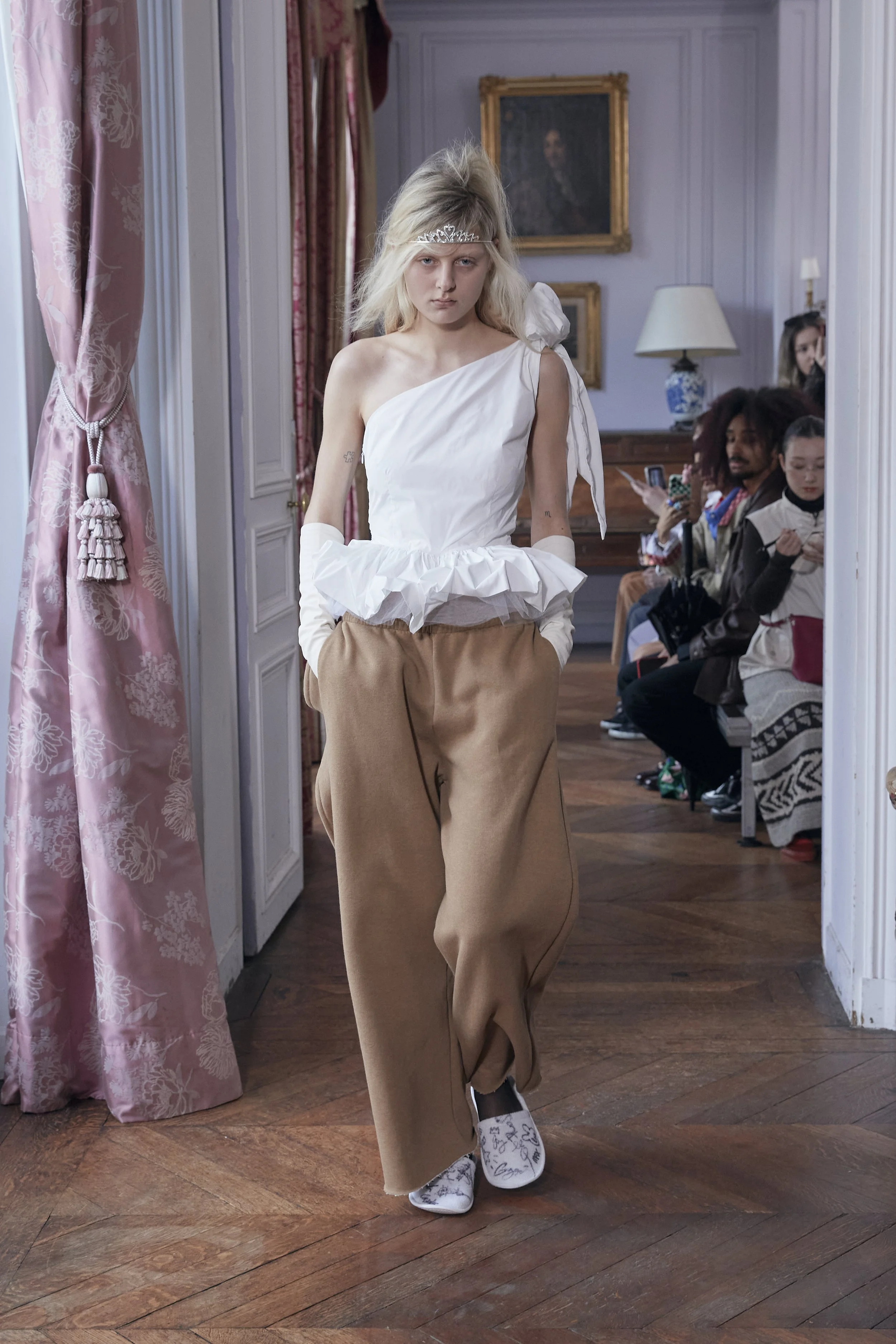 A young woman with blonde hair styled in a voluminous teased hairstyle wears a tiara, an asymmetrical white top with ruffled details, beige pants, and white shoes with black scribbles. She is walking in an elegant room with pink floral curtains, white paneled walls, classic paintings, and a group of spectators seated and watching her.