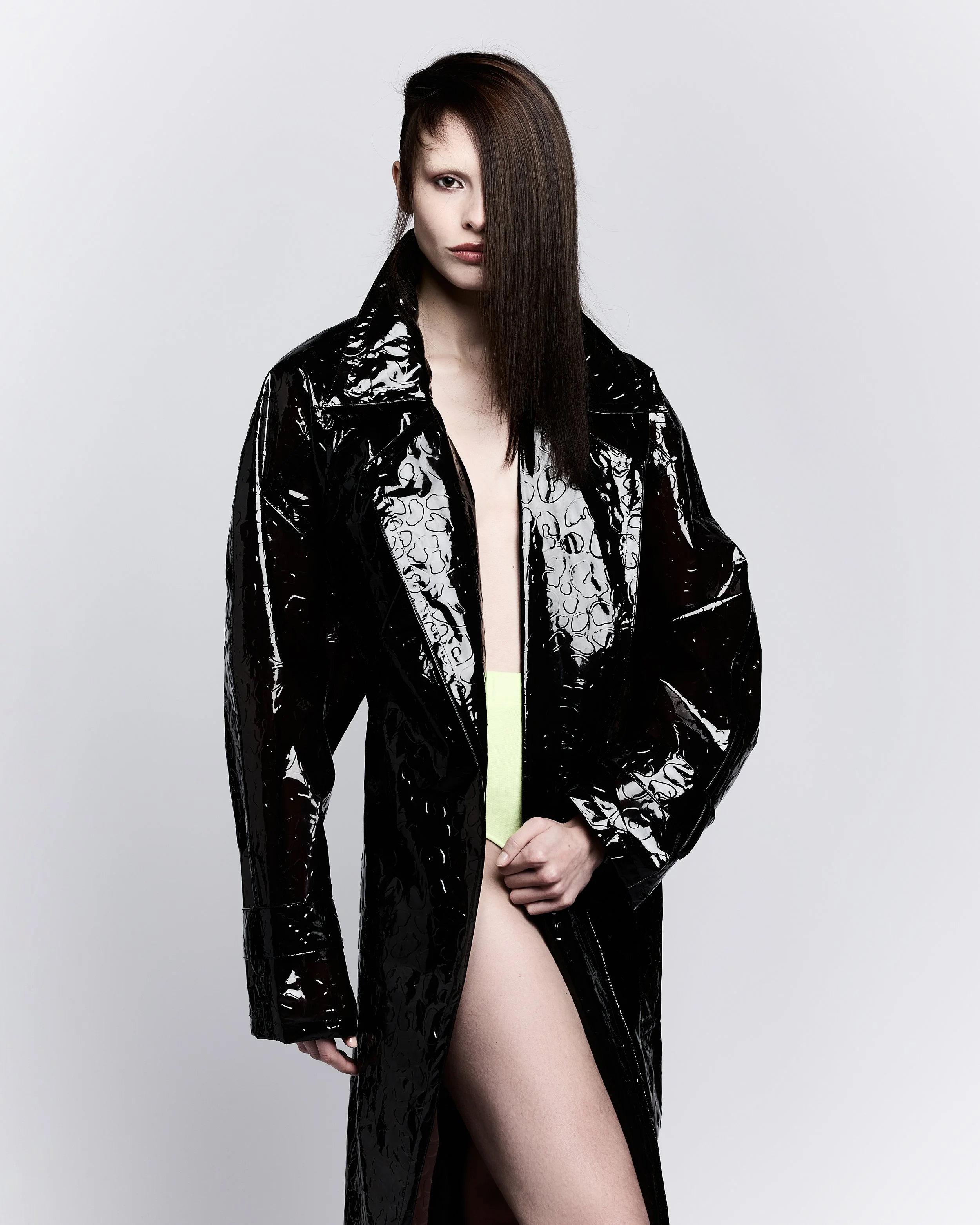 A woman with straight dark hair, half-covered face, wearing a black shiny coat with a leopard print lining, is standing against a plain light gray background, partially exposing her leg.