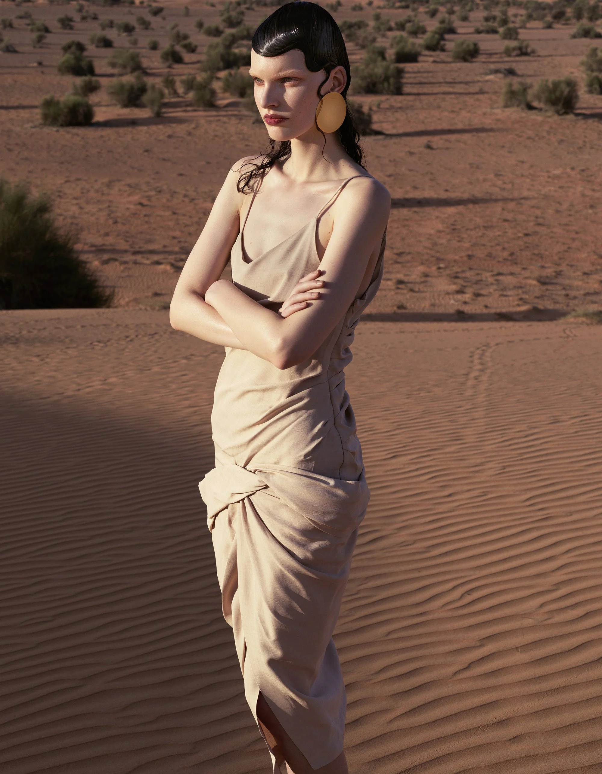 A woman with wet black hair styled in a vintage wave, wearing large gold earrings and a beige dress with spaghetti straps, standing in a desert landscape with sand dunes and sparse vegetation.