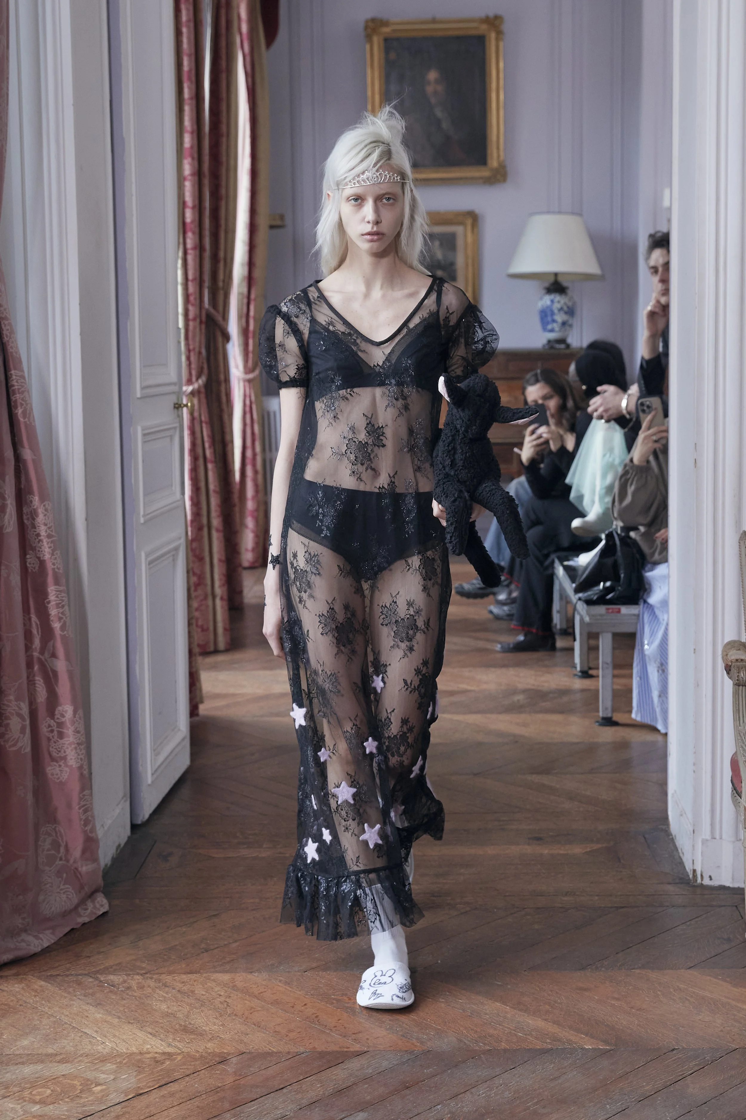 A young woman modeling black lace sheer clothing with star embellishments during a fashion show in an elegant room with hardwood floors, pink curtains, and framed artwork. She has platinum blonde hair, a silver headband, and white shoes with signatures or drawings on them. Several seated spectators are visible in the background.