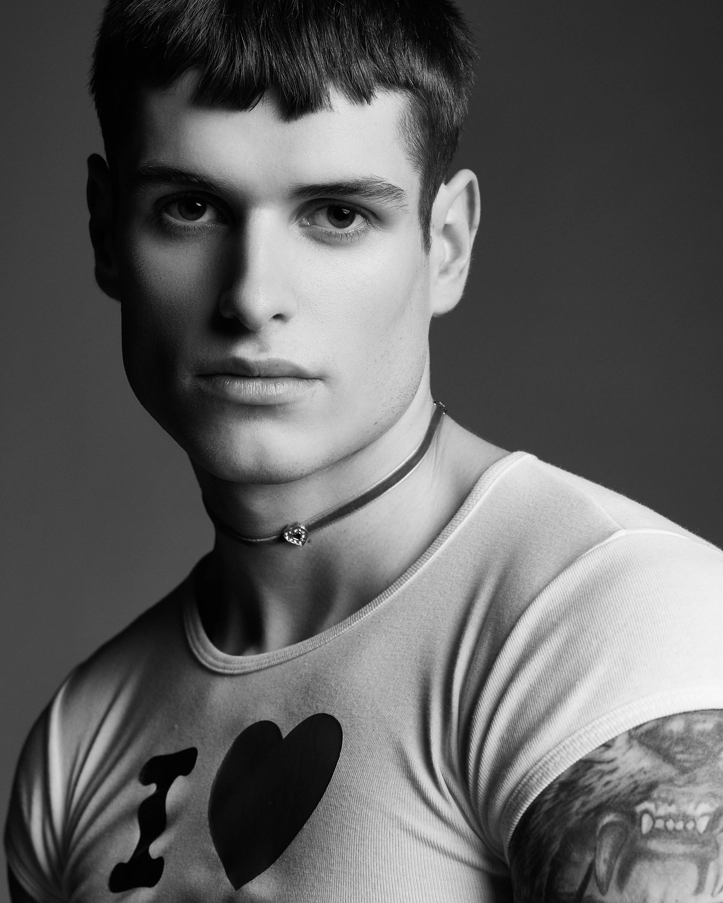Black and white photo of a person with light skin, short dark hair, tattoos on their arm, wearing a light-colored t-shirt with a large heart design and a choker necklace with a heart-shaped pendant.