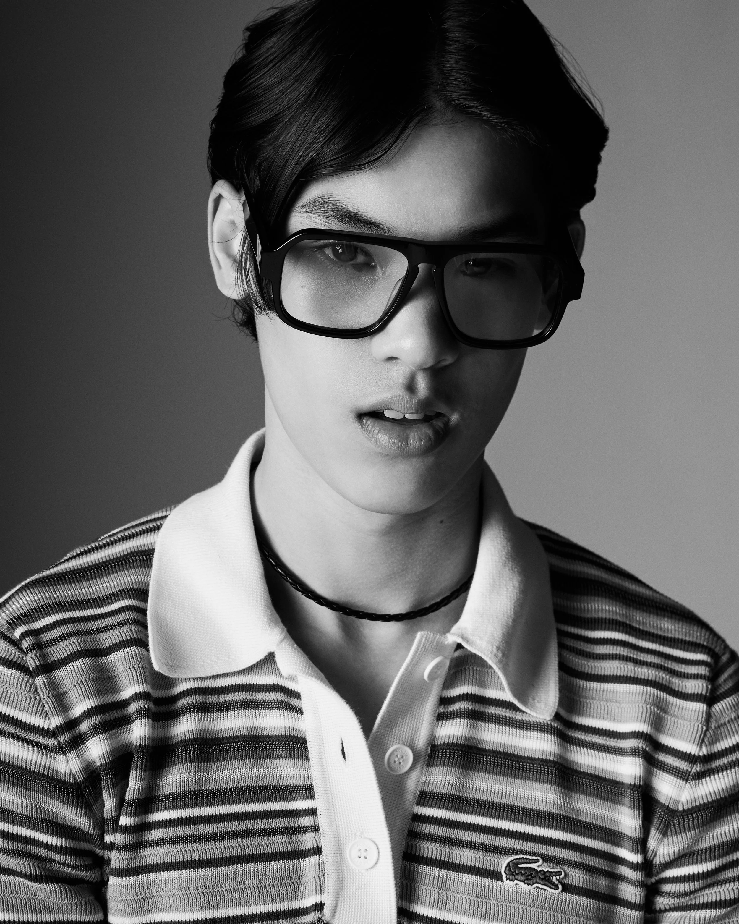 A black and white photo of a person with short dark hair, wearing large sunglasses, a collared shirt, a striped sweater, and a choker necklace.