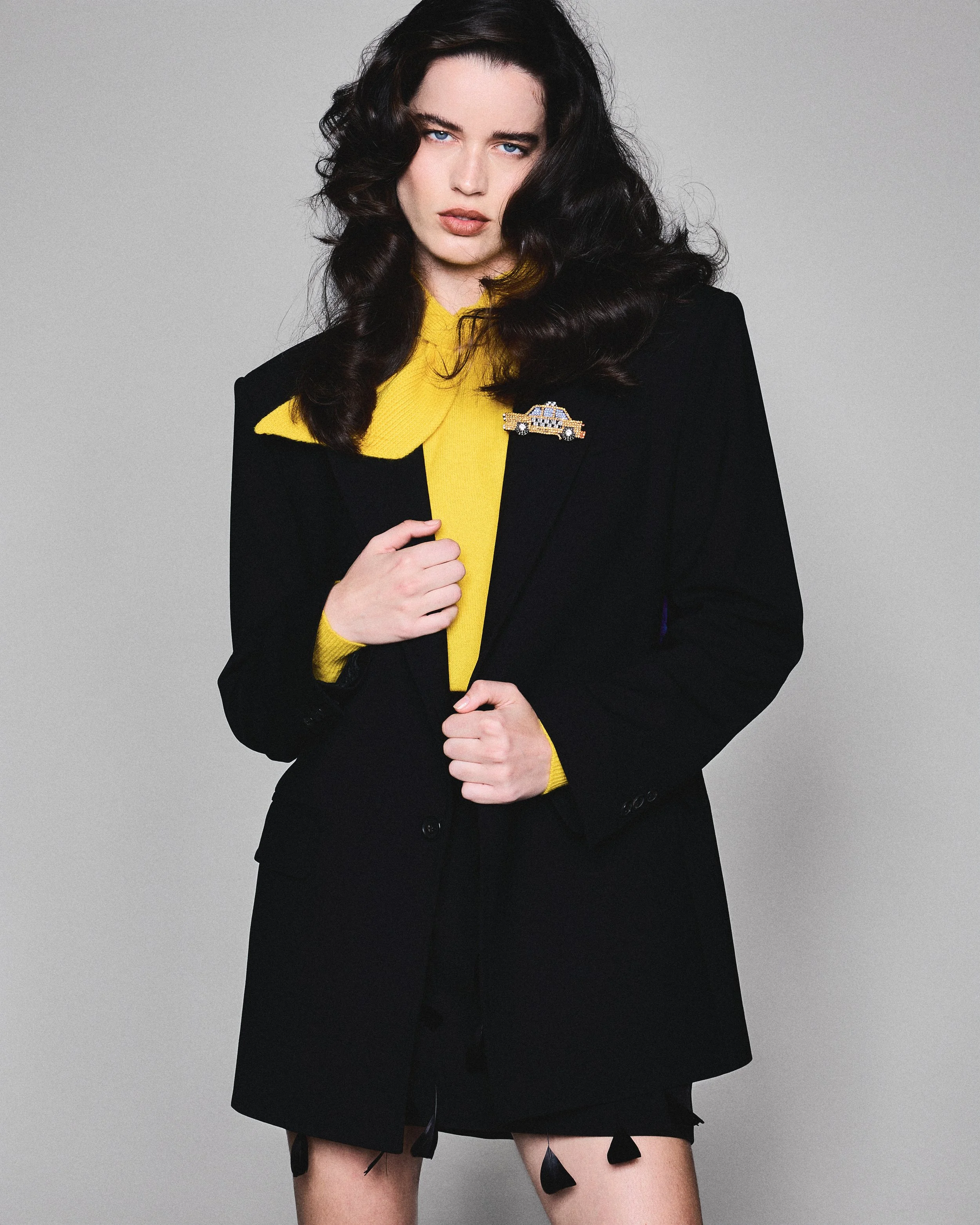 A woman with dark, wavy hair and blue eyes, dressed in a black blazer with a yellow shirt underneath, is holding the lapel of her blazer with one hand. She is wearing a police car brooch on her blazer and has torn, black ribbon-like pieces hanging from her blazer and skirt. The background is plain gray.