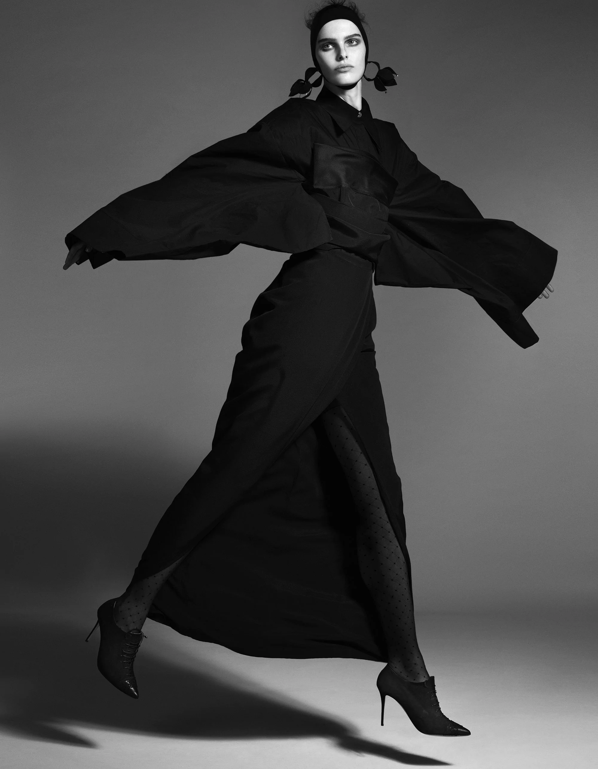 Black and white photo of a woman with dark clothing, a headband, and high-heeled boots in a dynamic pose with arms extended, against a plain gray background.
