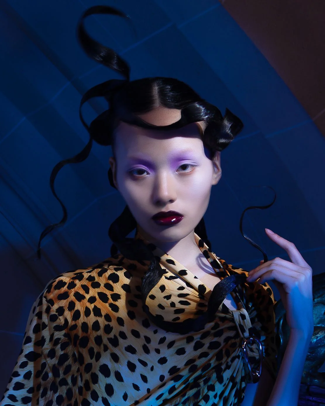 A woman with dark, glossy, curled hair, wearing leopard print clothing, has pale makeup with white and purple eyeshadow, and dark lipstick, in a dark, atmospheric setting.