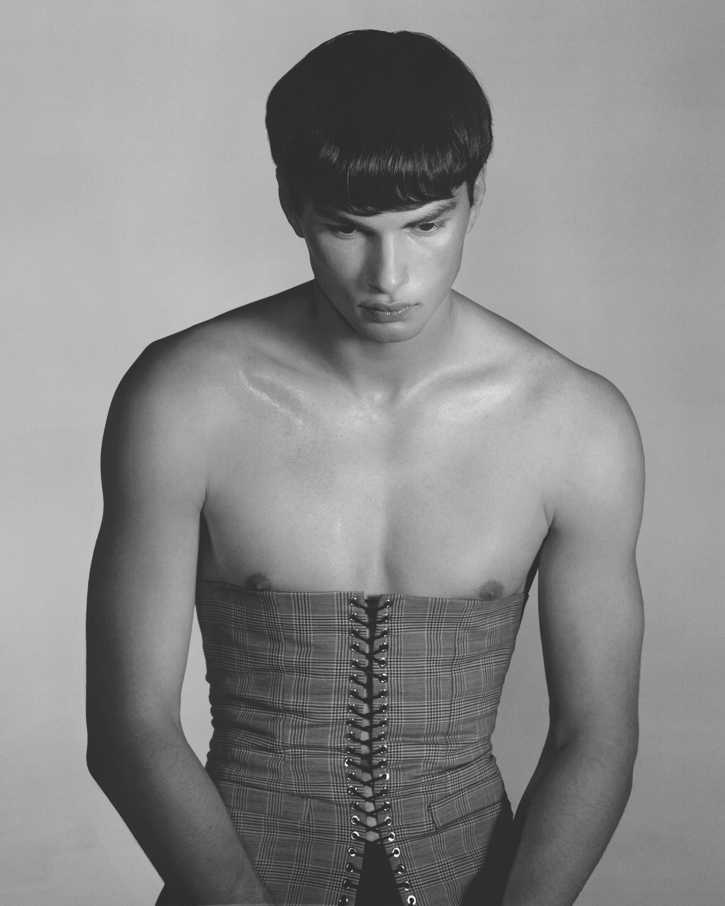 Black-and-white photograph of a young man with short hair, looking down. He is shirtless and wearing high-waisted plaid pants with a lace-up front.