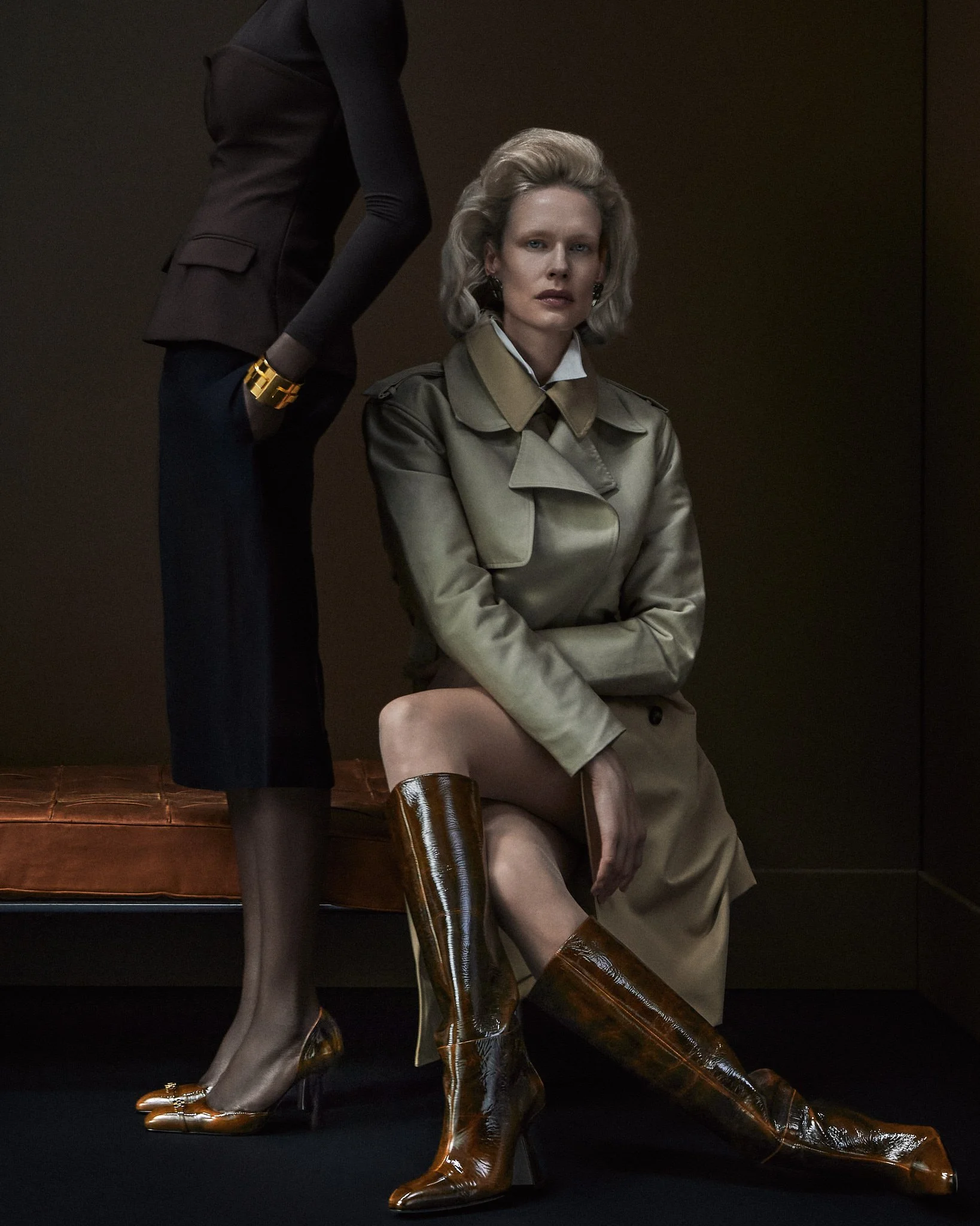 A woman with blonde hair, wearing a beige trench coat, tan pants, and tall shiny brown boots, sitting on a stool with her hands on her lap, and a person standing beside her with only part of their body visible, wearing a black blazer, dark pants, and a gold bracelet.