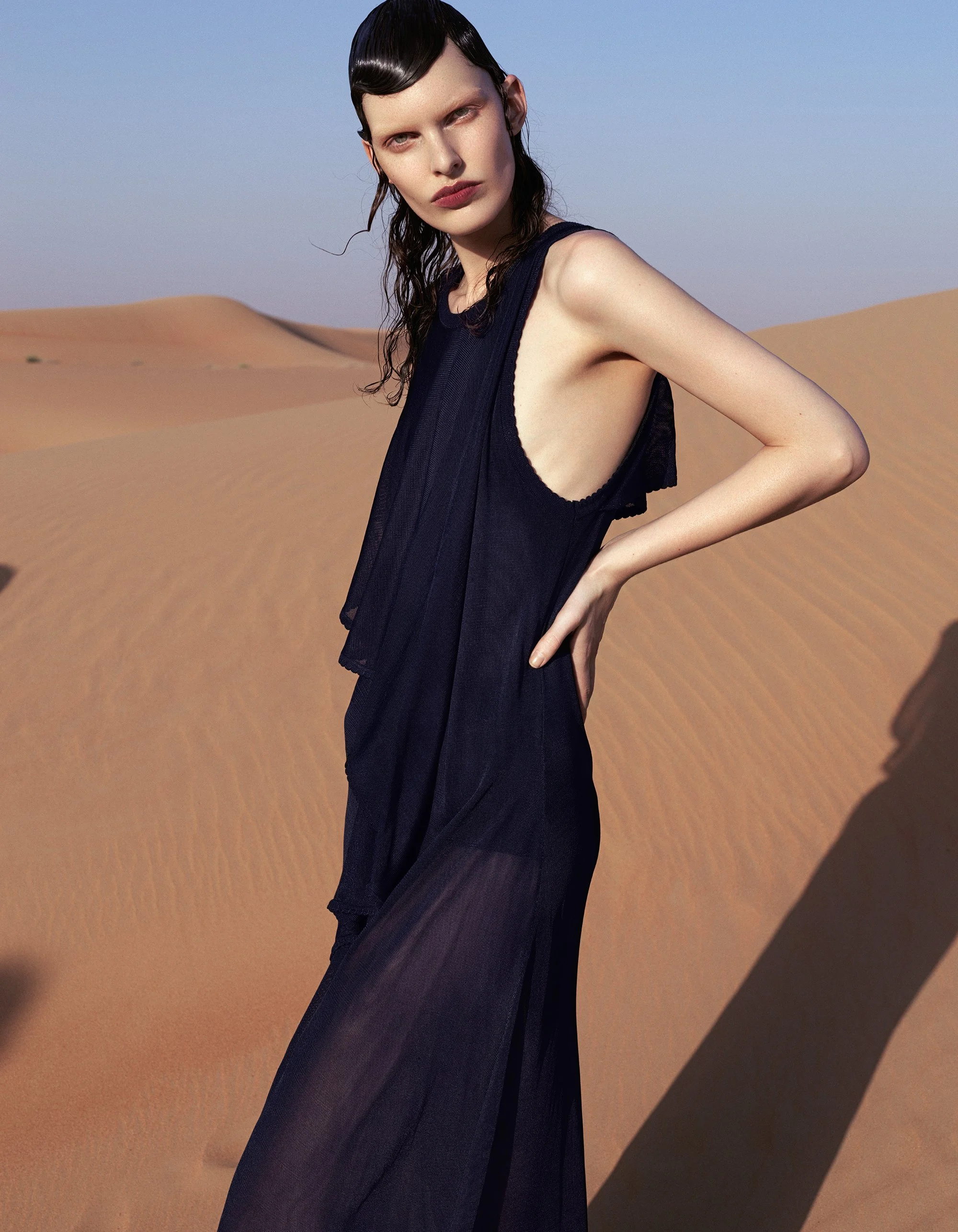 A woman with wet hair wearing a sleeveless black dress standing in a desert with sand dunes and a clear blue sky.