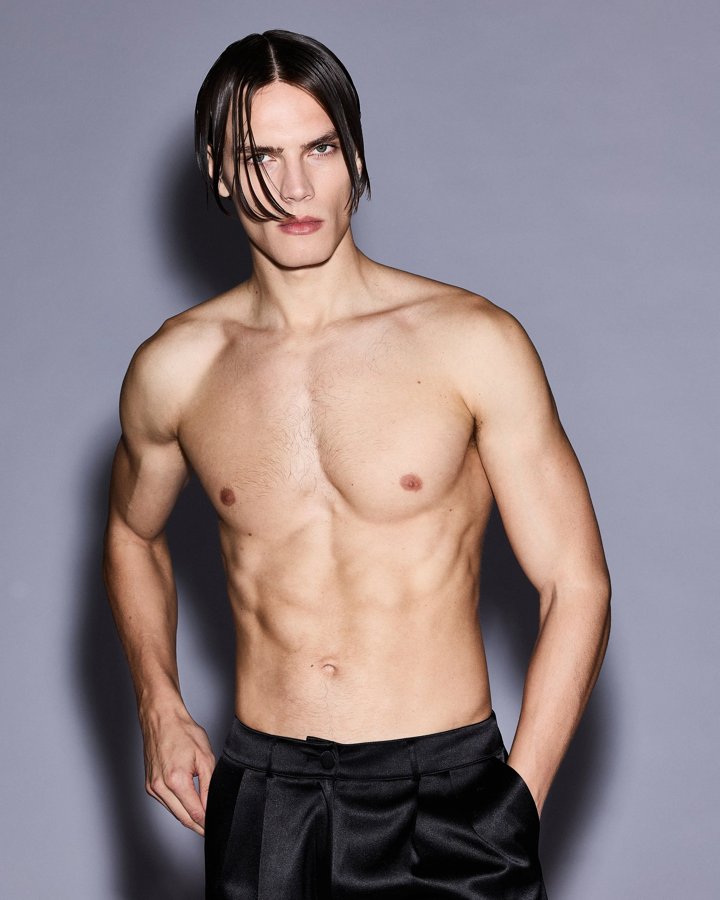 A shirtless man with dark hair styled to the side, standing against a neutral gray background, wearing black pants.