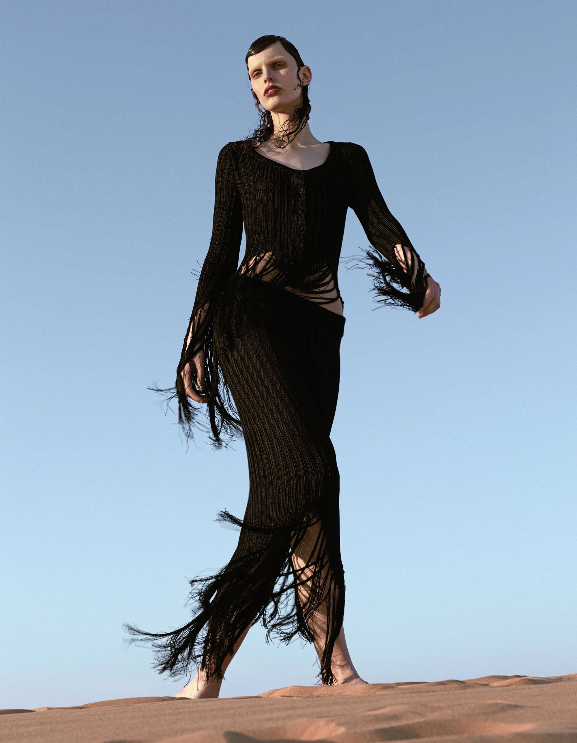 A woman standing on a sandy terrain with a clear blue sky in the background, wearing a long, black, sheer, fringed dress.