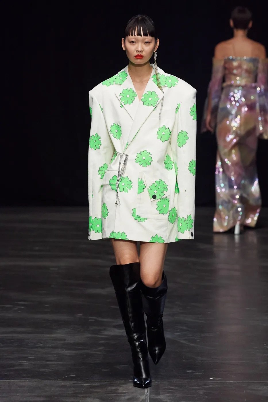 Model walking on runway wearing an oversized white blazer with green flower patterns, paired with black knee-high boots, during a fashion show.