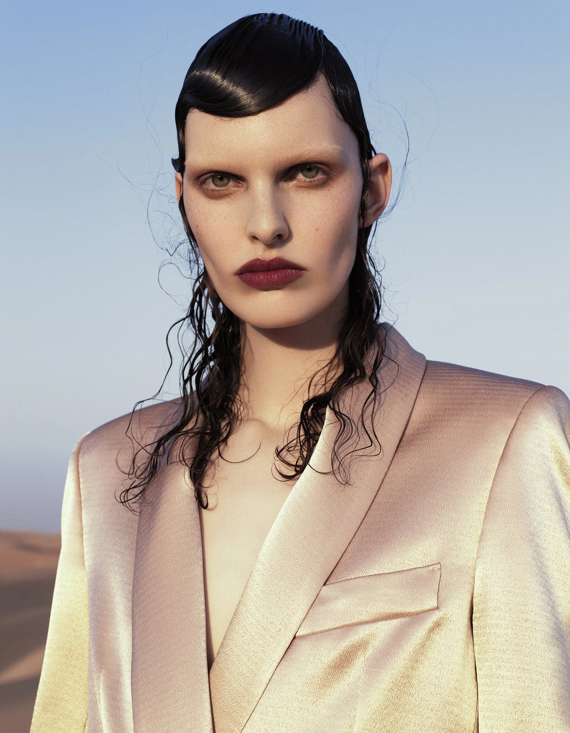 Close-up of a woman with wet black hair, wearing a beige blazer, against a soft outdoor background.