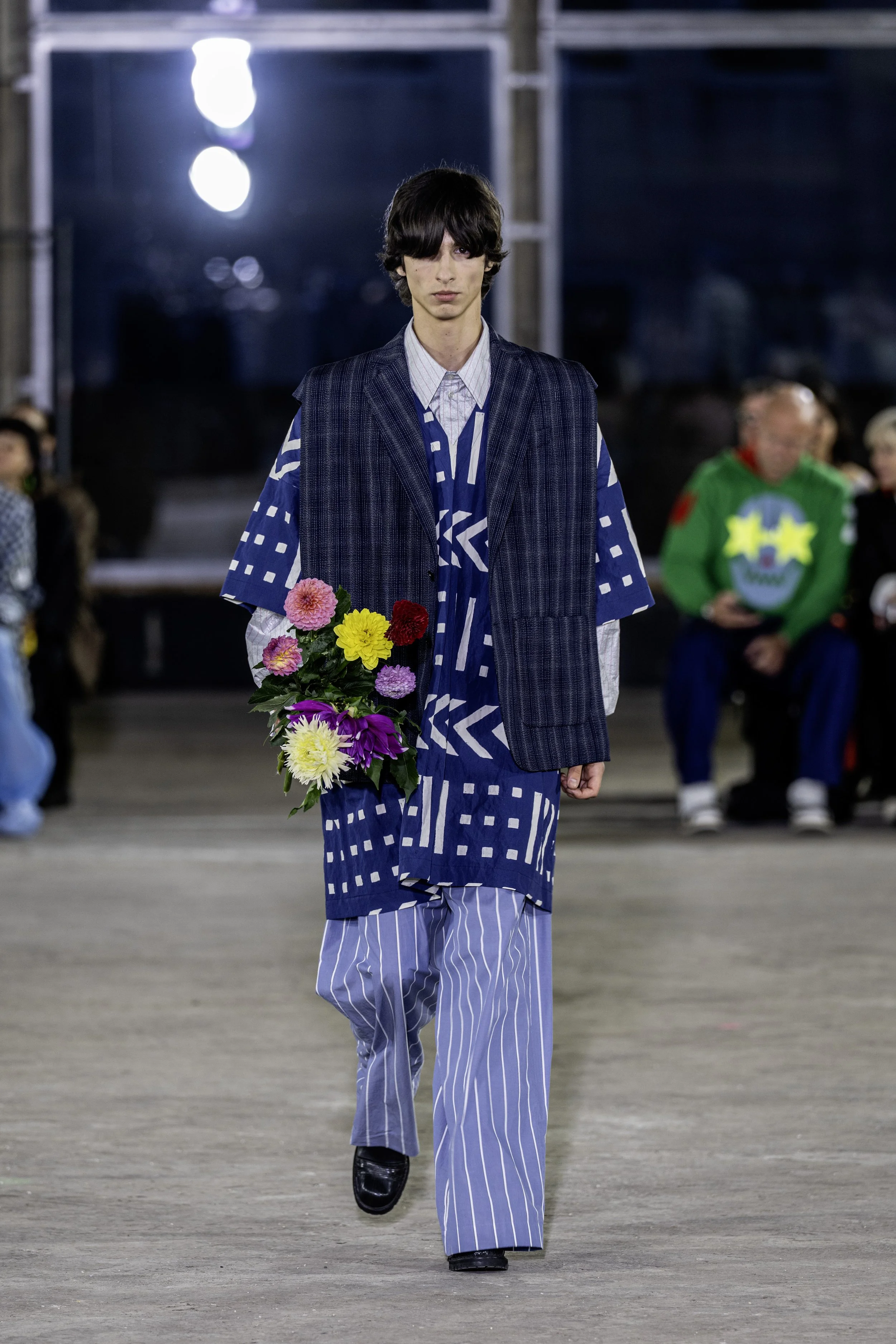 A runway model in a fashion show holds a bouquet of multicolored flowers, wearing a layered outfit with blue and white patterns, and walking on a runway with an audience in the background.