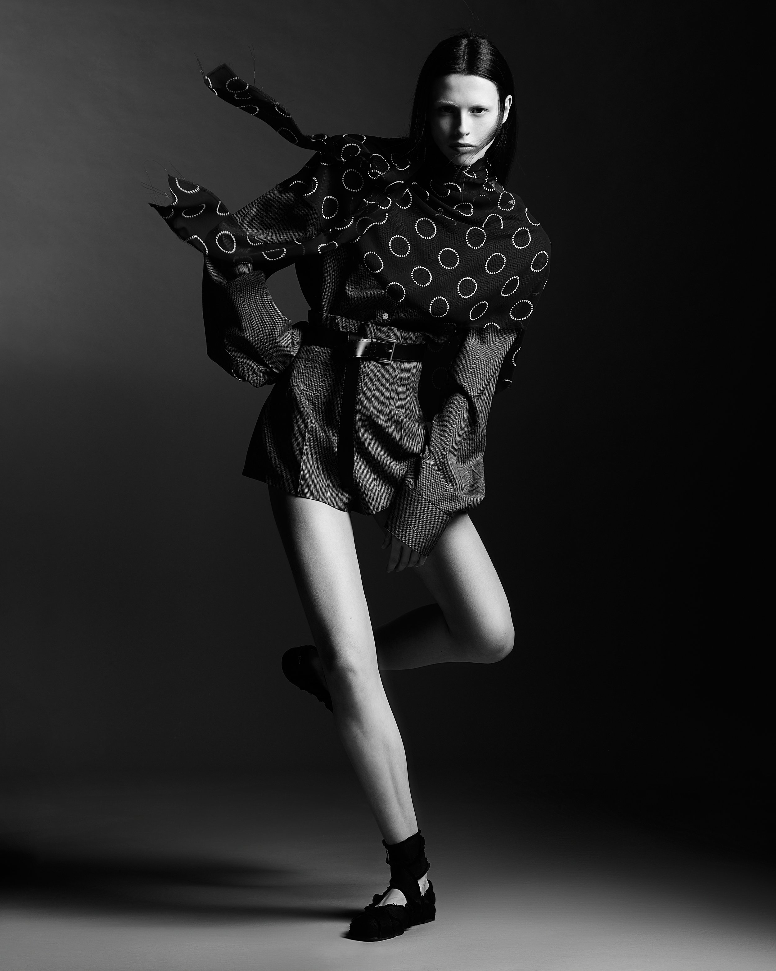 Black and white fashion photo of a woman in a polka dot blouse, high-waisted shorts, and sneakers, posing with one leg bent and her hand resting on her knee against a gradient background.
