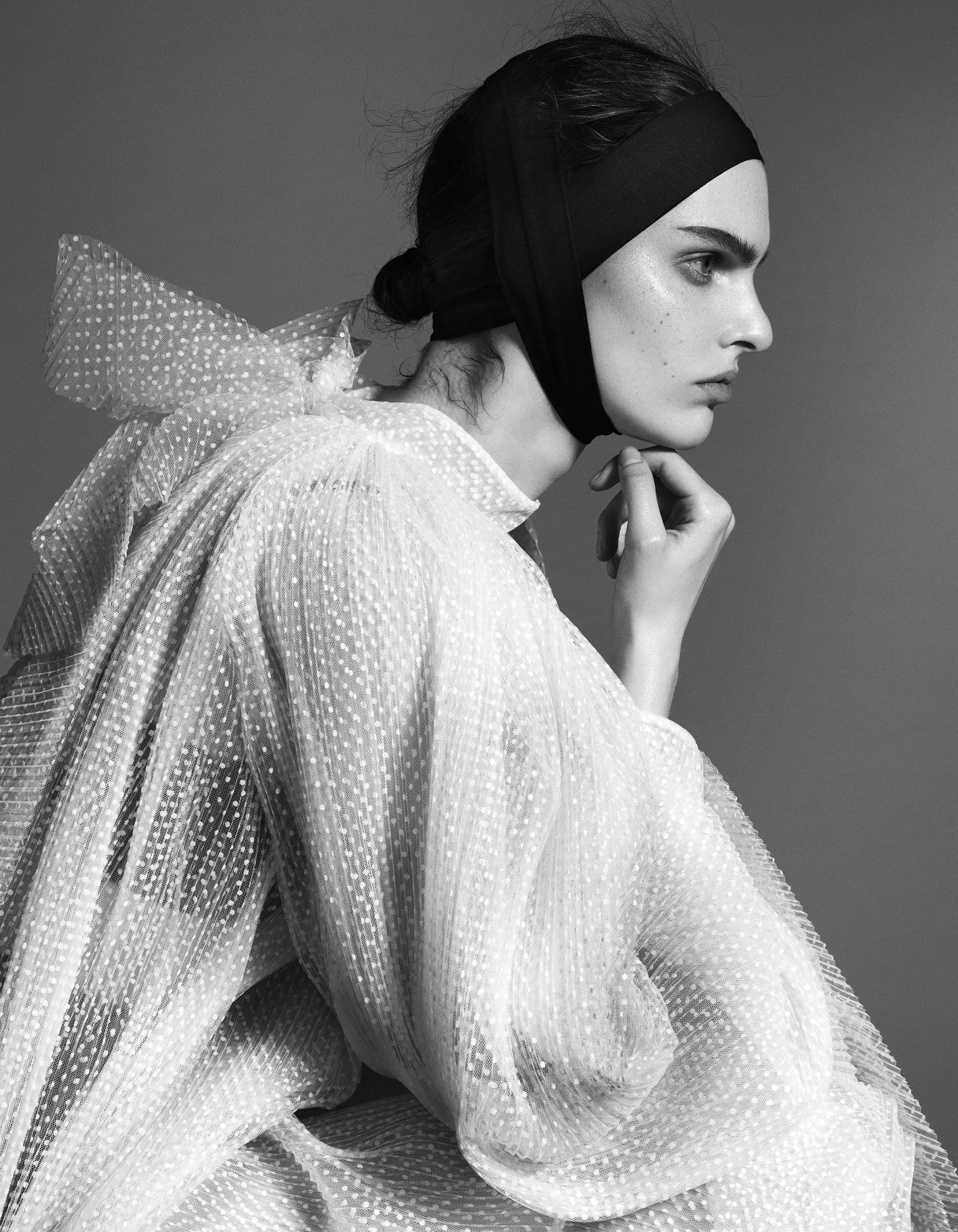 Black and white photo of a woman with dark hair, wearing a black headband, looking to the side with a serious expression. She is dressed in a textured, ruffled, semi-transparent fabric with small dots.