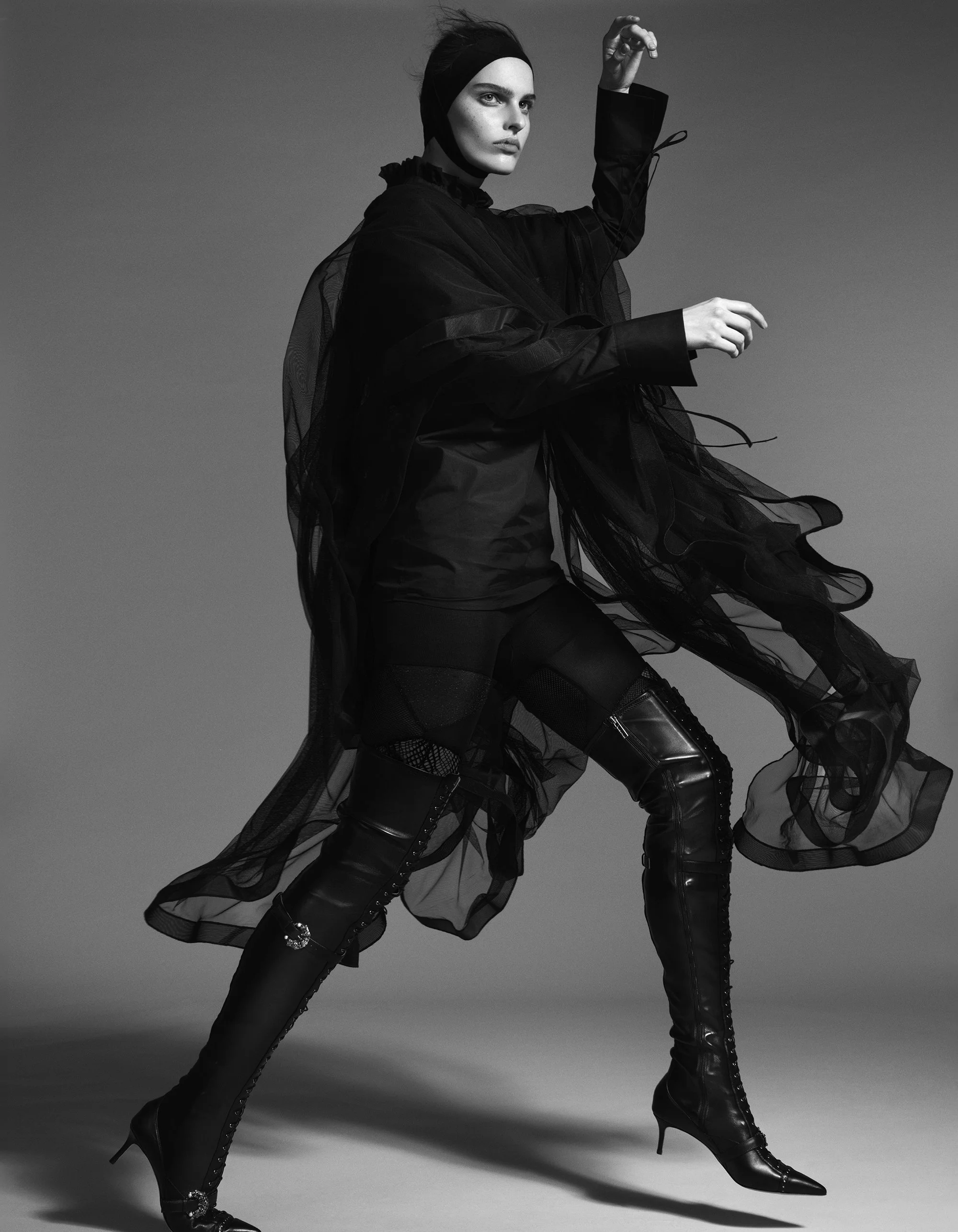 Black and white photo of a woman in fashionable clothing posing against a plain background. She wears a headband, a sheer ruffled cape, dark leggings, and high-heeled knee-high boots, with one arm raised and the other cast downward.
