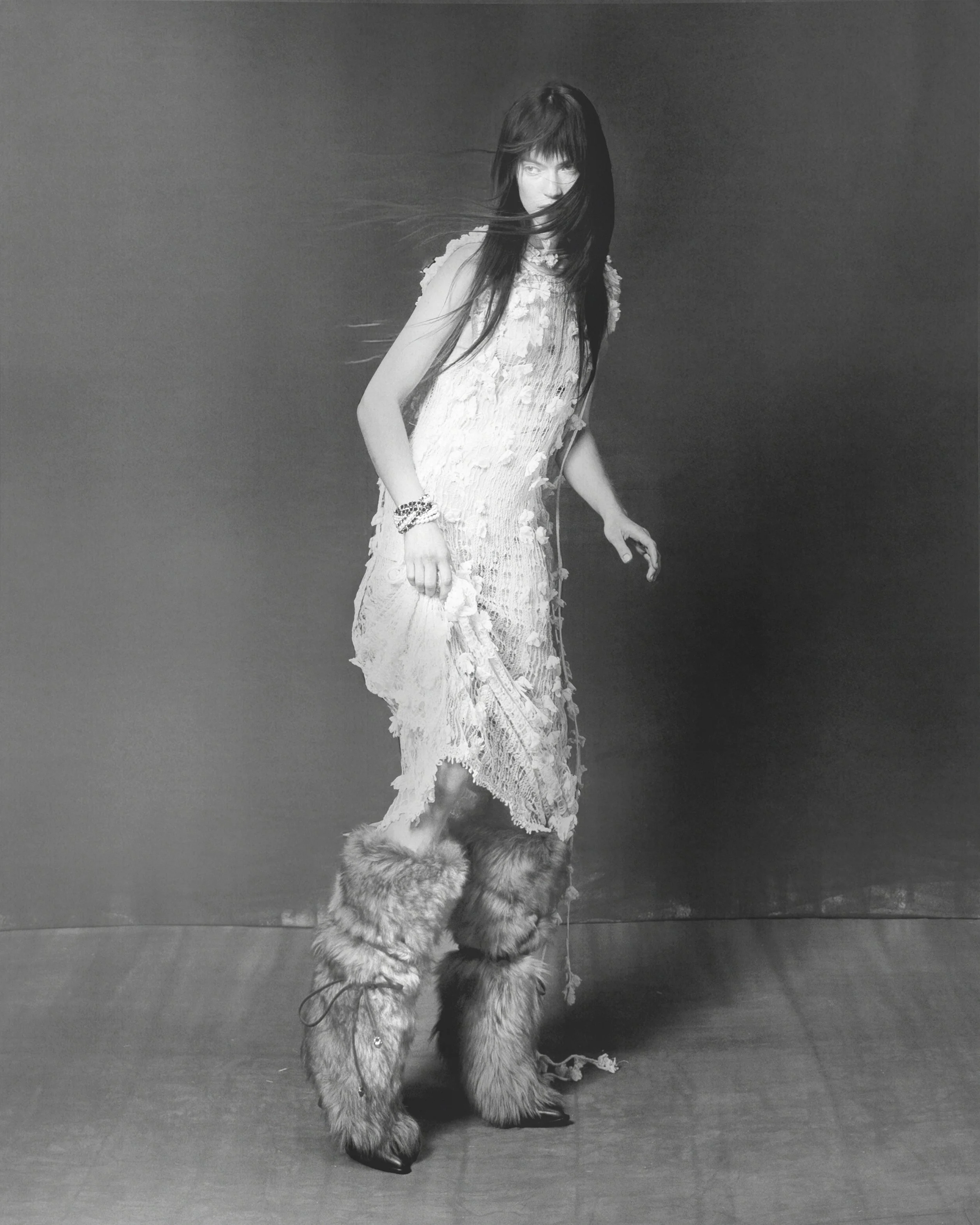 A woman with long dark hair, wearing a lace dress and fur boots, stands on a plain background, with her hair blowing across her face.