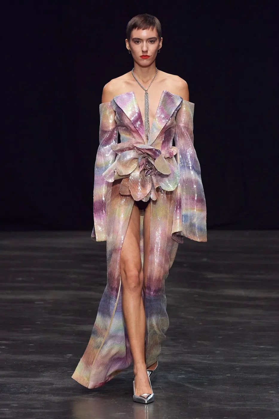Model walking on runway wearing iridescent, off-the-shoulder dress with floral design and high slit, paired with silver high heels, dark background.