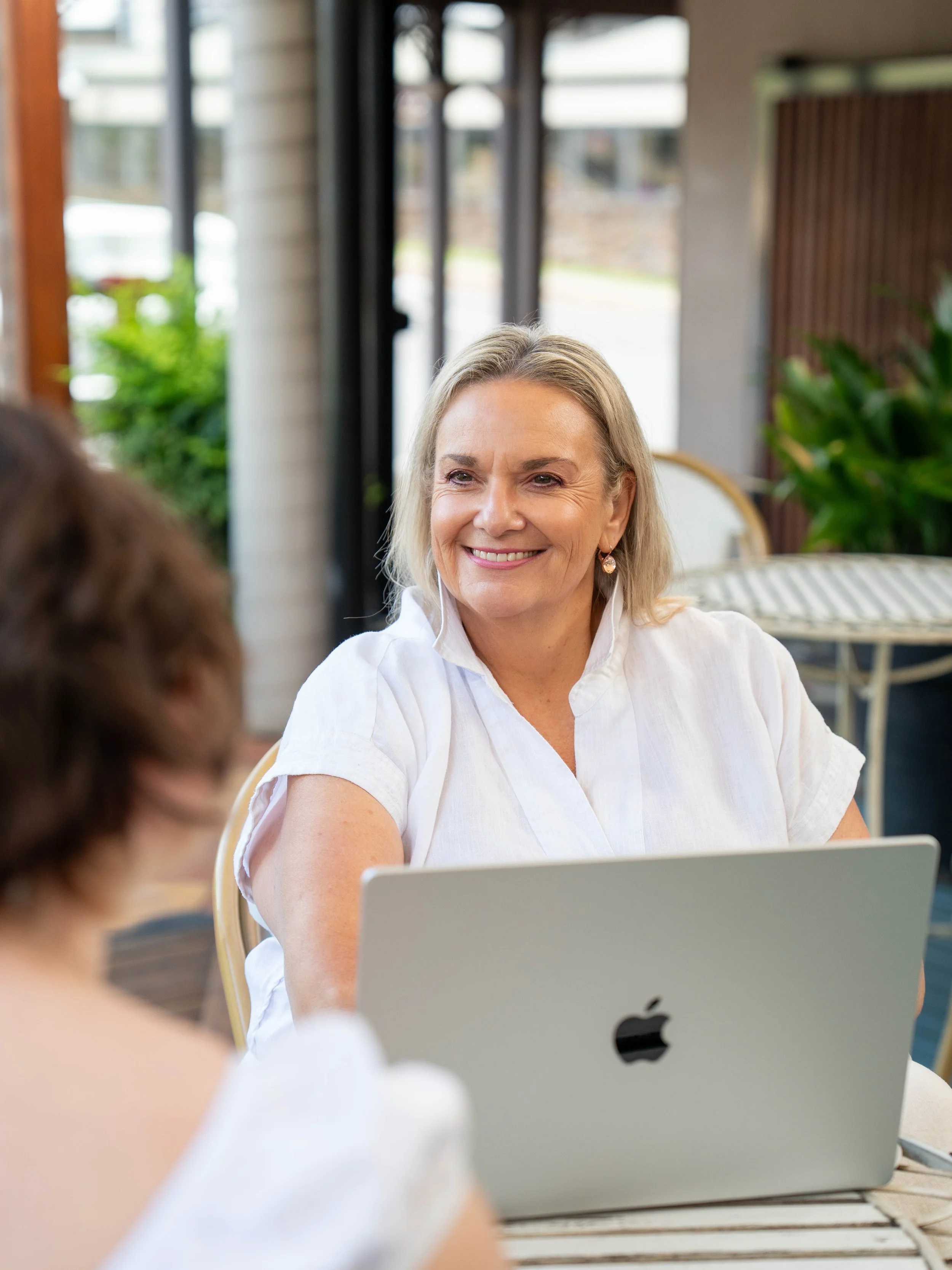 Smiling woman in a white blouse using an Apple laptop indoors, assisting an elderly person blurred in the foreground
