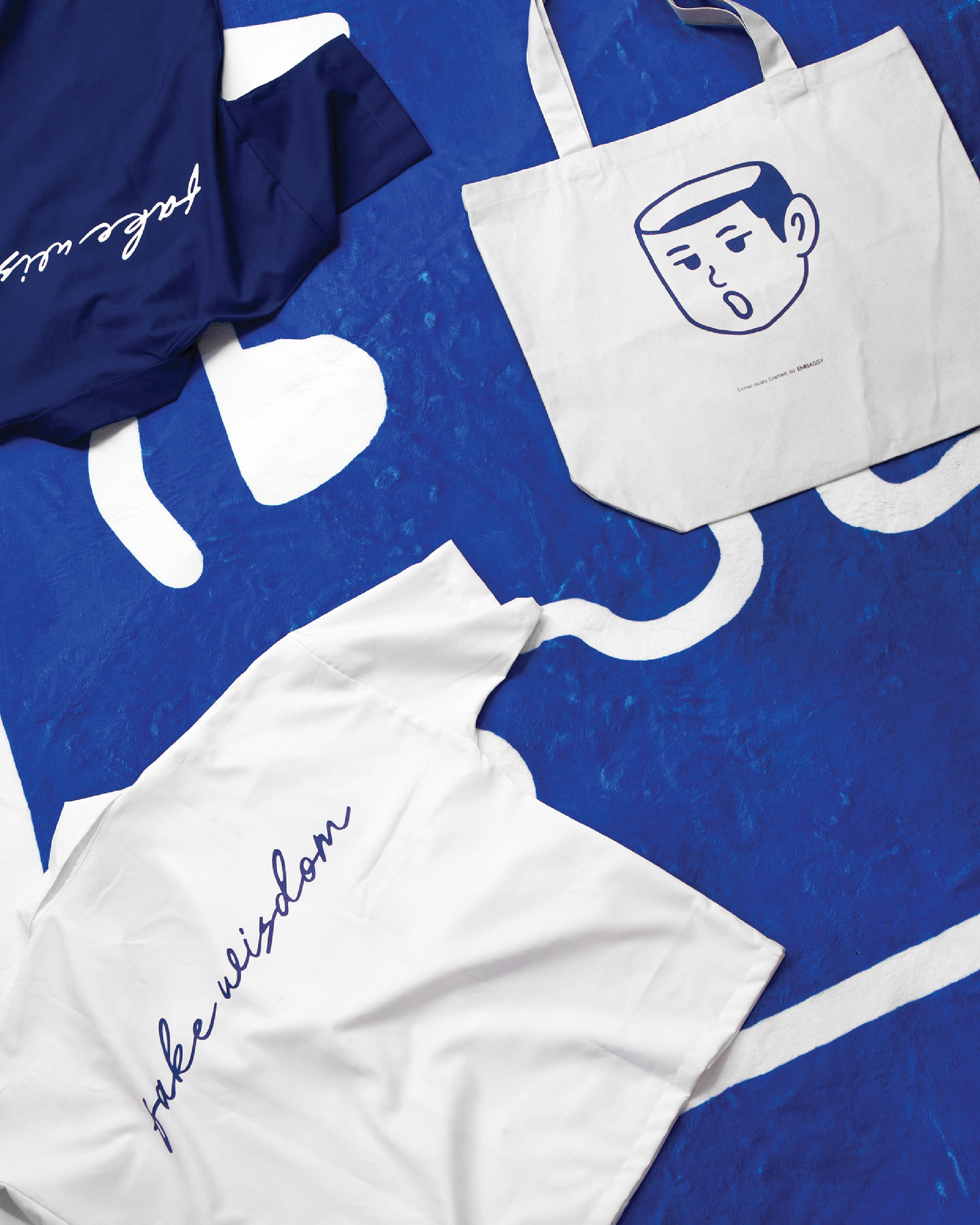 A white tote bag with a blue outline drawing of a person's face, lying on a blue surface with white design elements. The tote bag has text that says 'the unquestioned' in blue cursive.