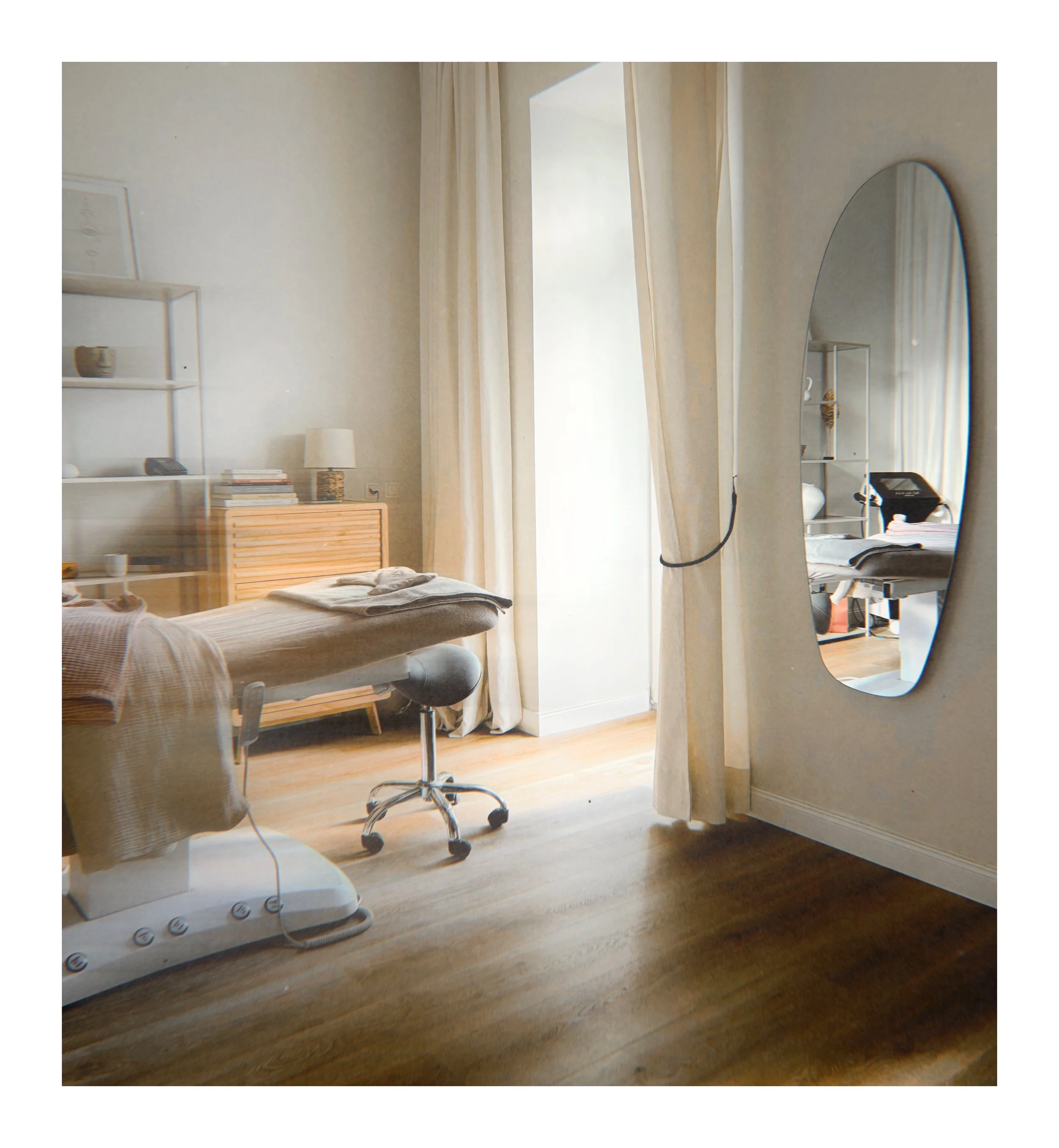 A cozy bedroom with a bed covered in neutral-colored bedding, a bedside table with a lamp and a stack of books, a large mirror reflecting part of the room, and a window with curtains allowing natural light into the space.