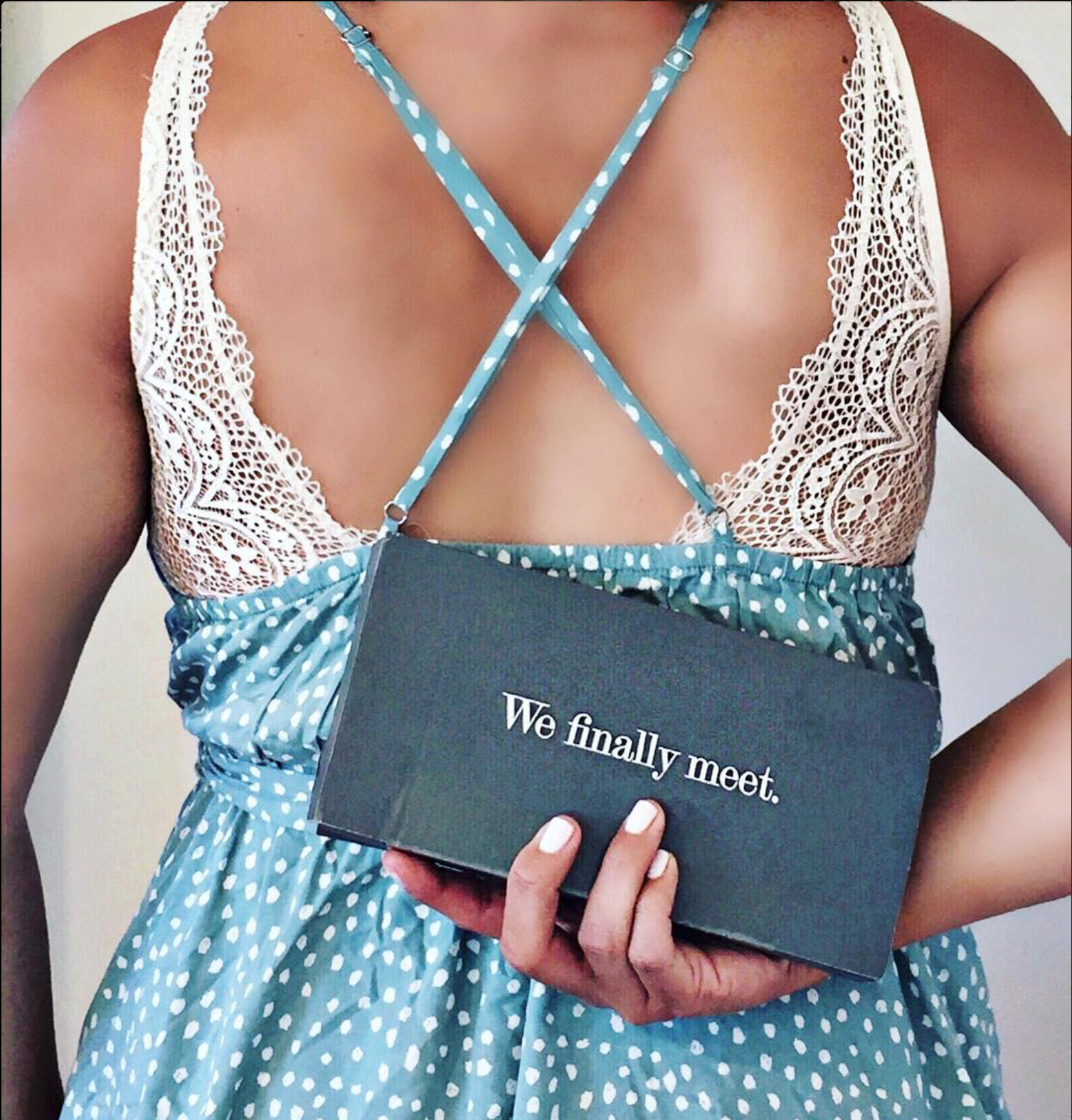 A woman wearing a blue dress with white polka dots and lace details, holding a black box that says 'We finally meet.'
