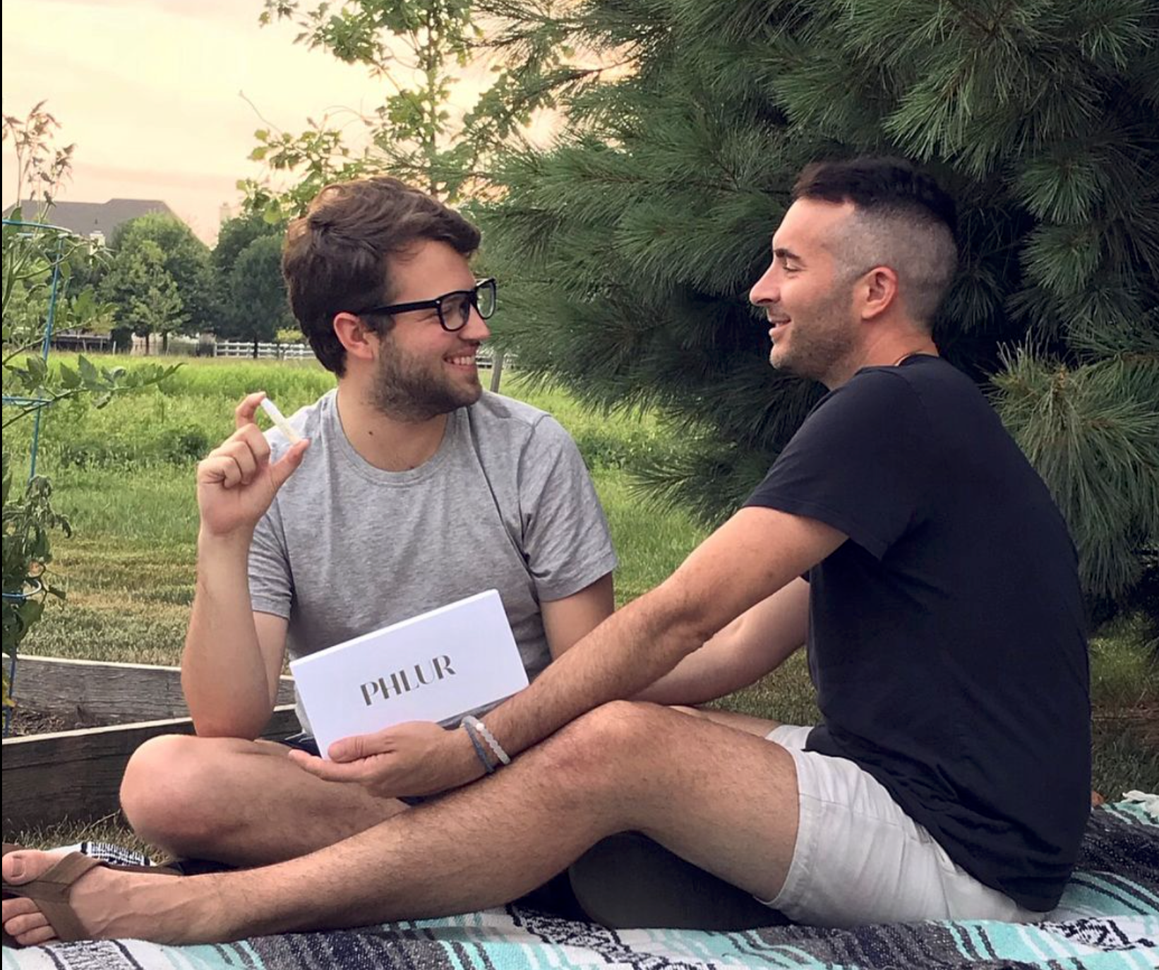 Two men are sitting on a blanket outdoors, smiling and looking at each other. One man has glasses, a gray t-shirt, and is holding a cigarette, while the other man is wearing a black t-shirt and white shorts. There is a sign with the name 'PHLUR' on his lap. They are seated near a pine tree, with a grassy field and trees in the background during sunset.