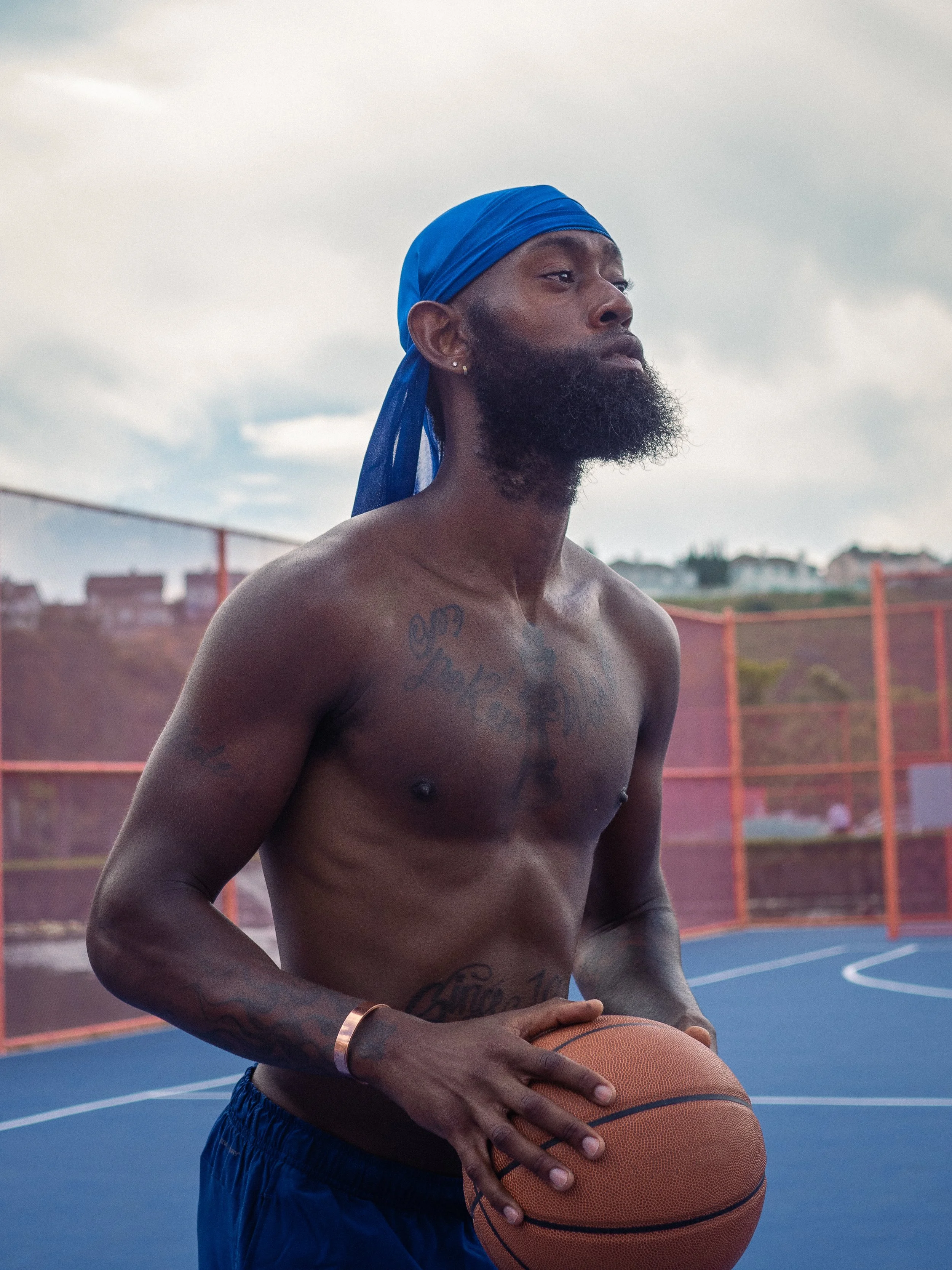 A shirtless man with a beard, wearing a blue headscarf and blue shorts, holding a basketball on an outdoor basketball court under a cloudy sky.