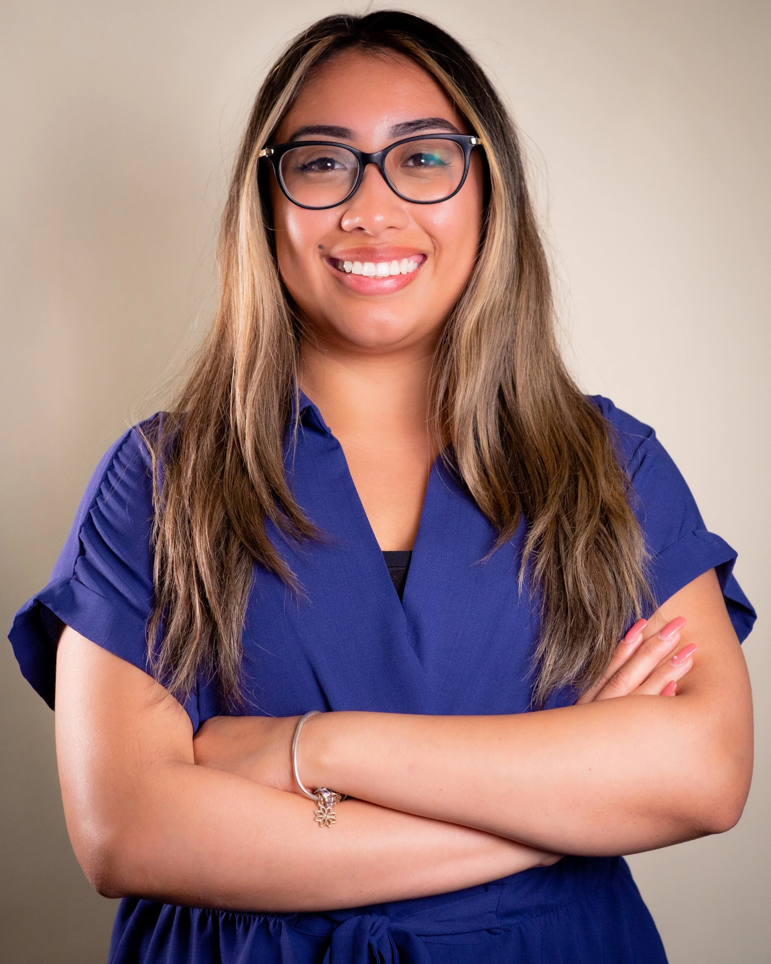 Young woman with long brown hair, glasses, smiling, wearing a blue dress with arms crossed.