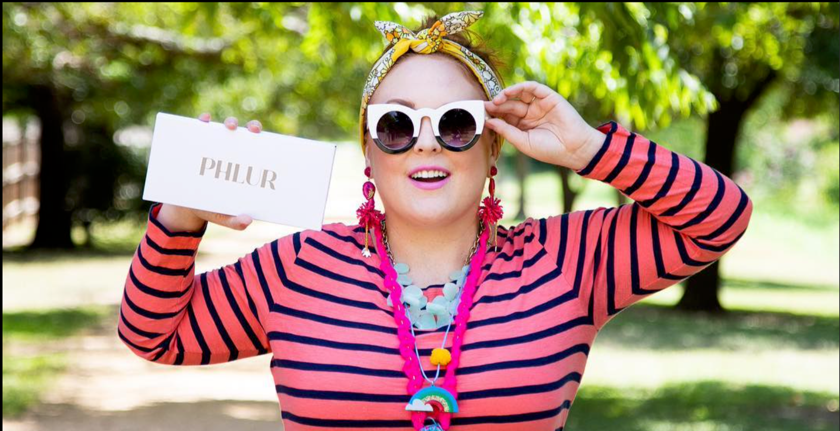 A woman outdoors holding a white sign wearing large round sunglasses, a yellow headscarf, a striped pink and blue shirt, and colorful jewelry, smiling at the camera.