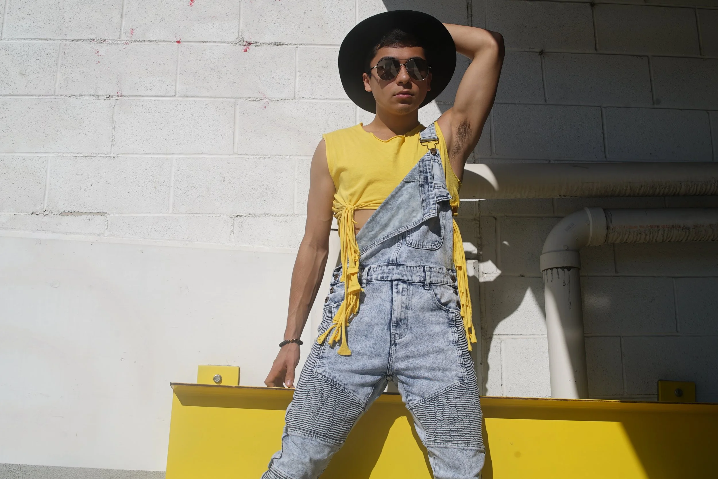 Person wearing a yellow sleeveless top, denim overalls, wide-brimmed black hat, and sunglasses, standing against a white brick wall with yellow background and pipes.