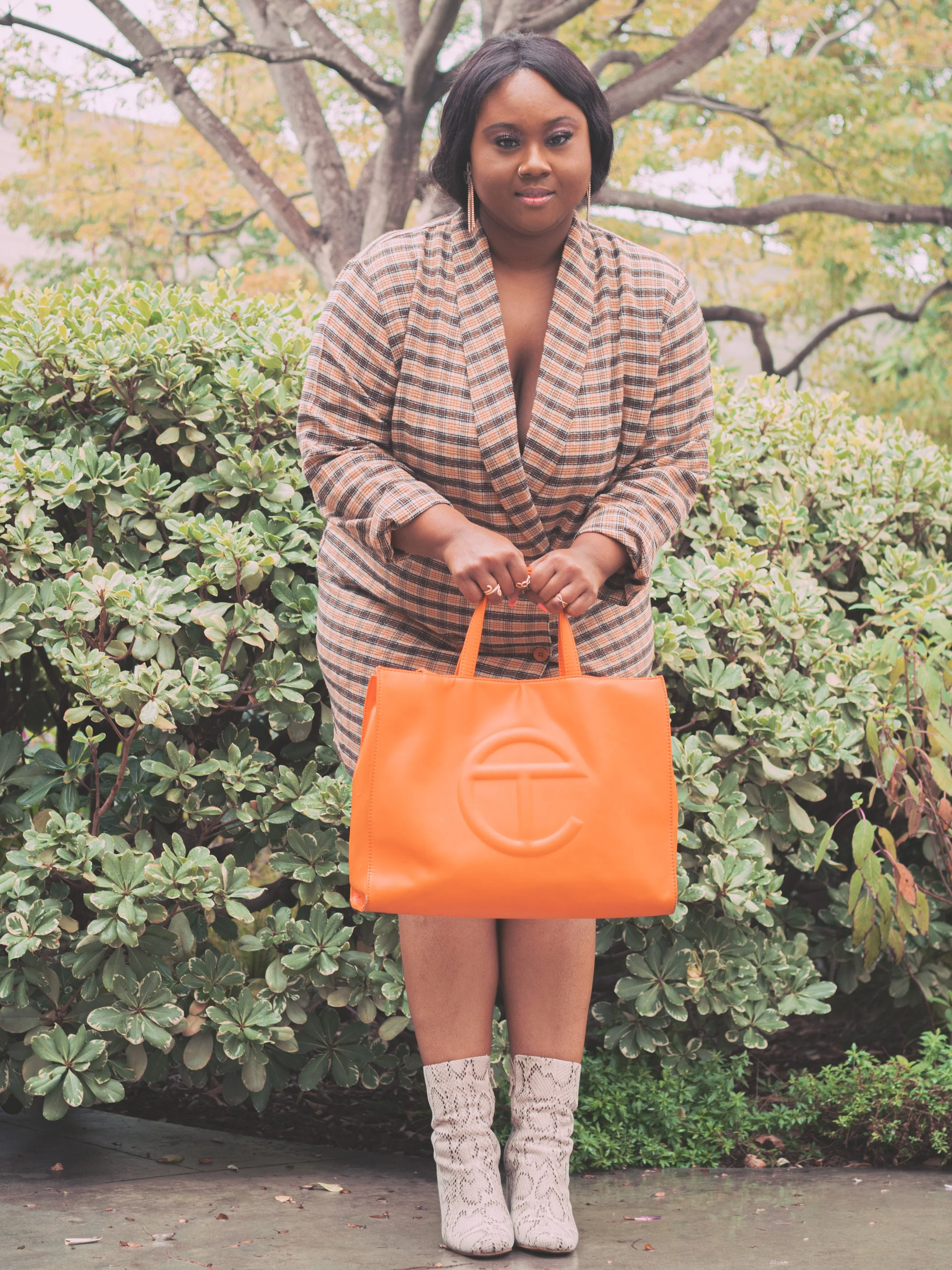 Woman in plaid blazer holding an orange tote bag with a large T logo, standing outdoors in front of green shrubs and trees, wearing white snakeskin boots.