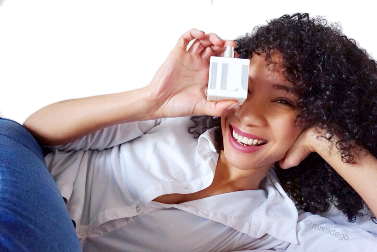A woman with curly dark hair and a white shirt lying on her side, smiling while holding a perfume bottle up to her eye.
