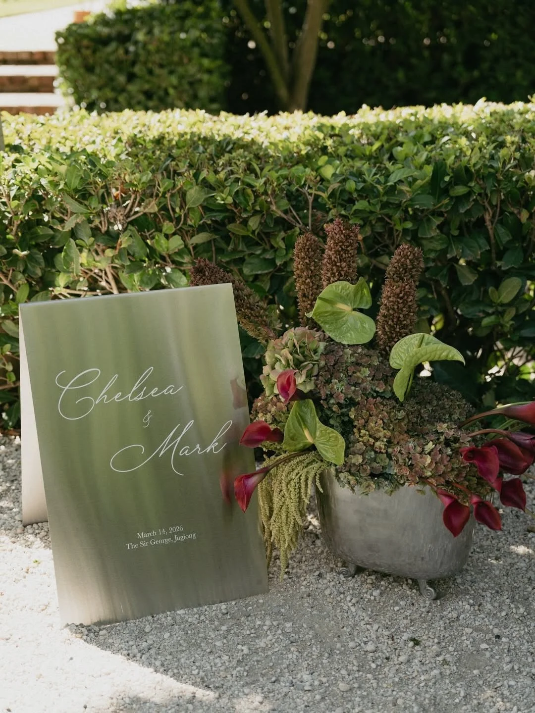 Wedding sign with names Chelsea and Mark, date March 14, 2026, at The Sir George, Jugiong, placed next to a floral arrangement in a silver planter outdoors with a background hedge.