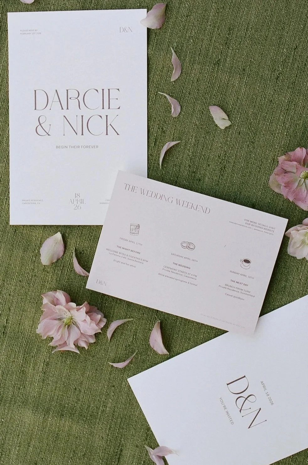 Wedding invitation suite with pink flower petals on green textured fabric. Includes a large invitation for Darcie & Nick, a schedule of events for the wedding weekend, and a smaller invitation with the name 'D&N' and the date April 14, 2023.