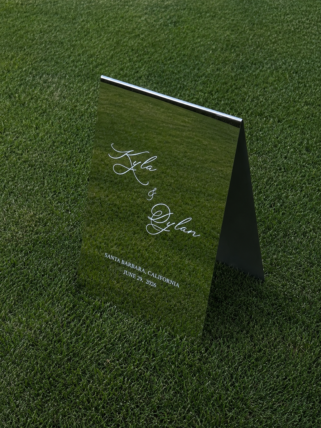 A reflective wedding sign with cursive text on a grassy surface. The sign reads ''Kyla & Dylan,'' with the location 'Santa Barbara, California' and date 'June 29, 2026' written at the bottom.