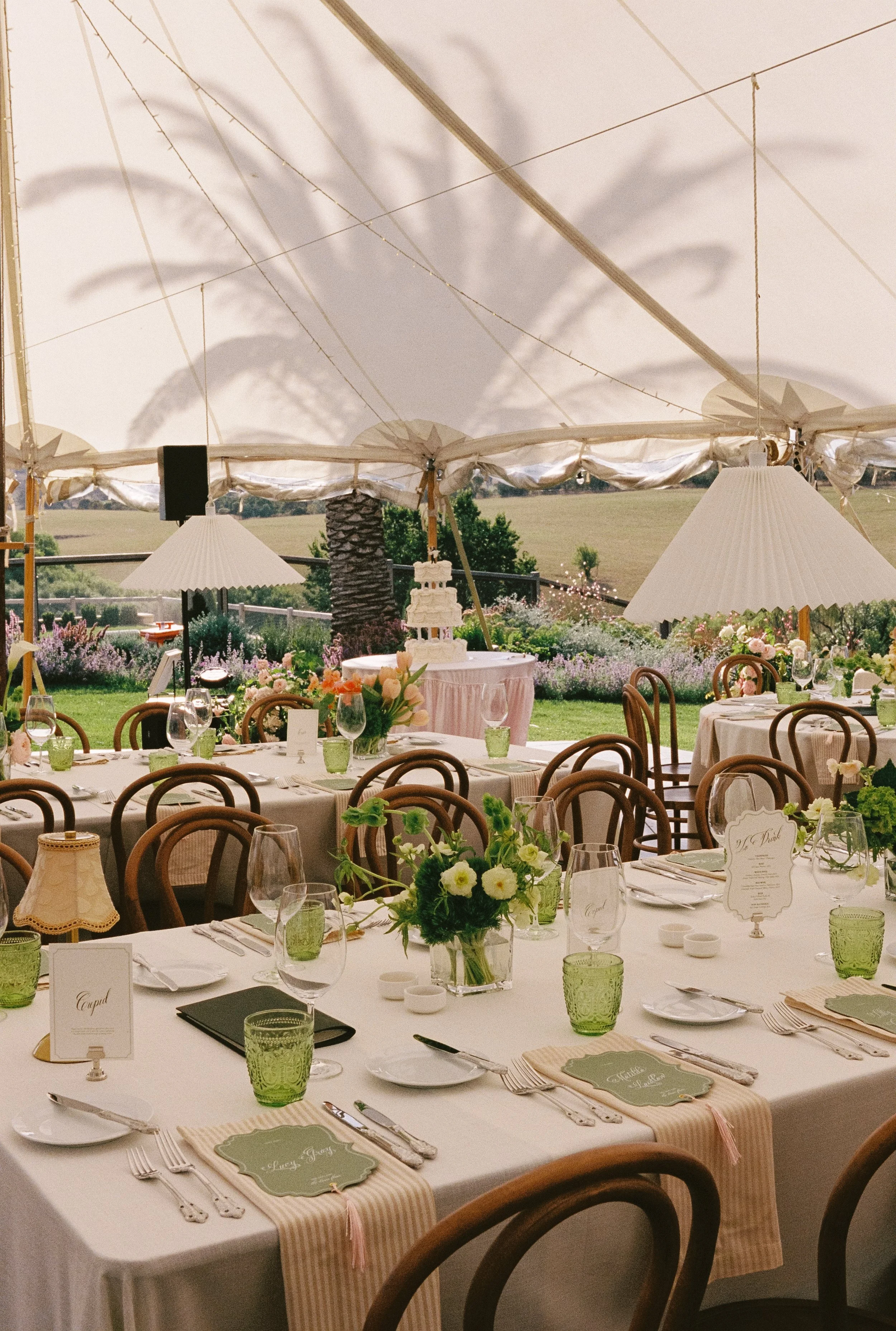 Elegant outdoor wedding reception setup under a large white tent with decorated tables, floral centerpieces, and place cards, overlooking a lush green landscape.