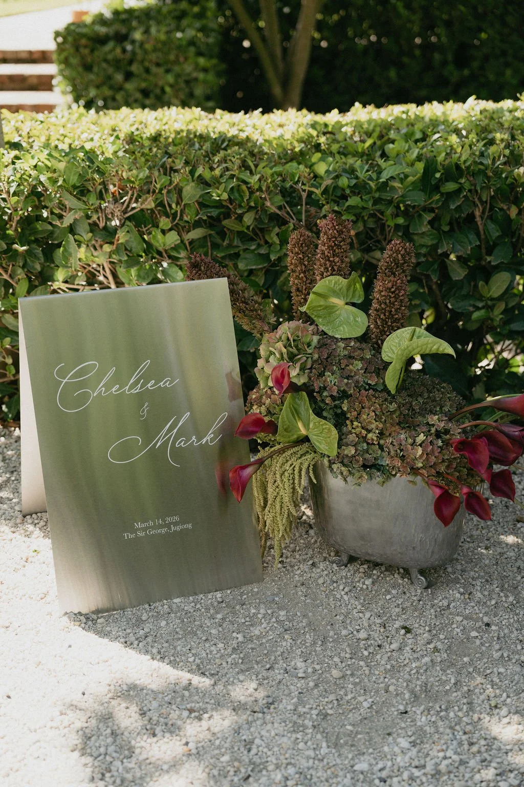 Wedding sign with the names Chelsea and Mark, dated March 14, 2026, placed outdoors next to a flower arrangement with green and purple foliage, in front of a hedge and steps.