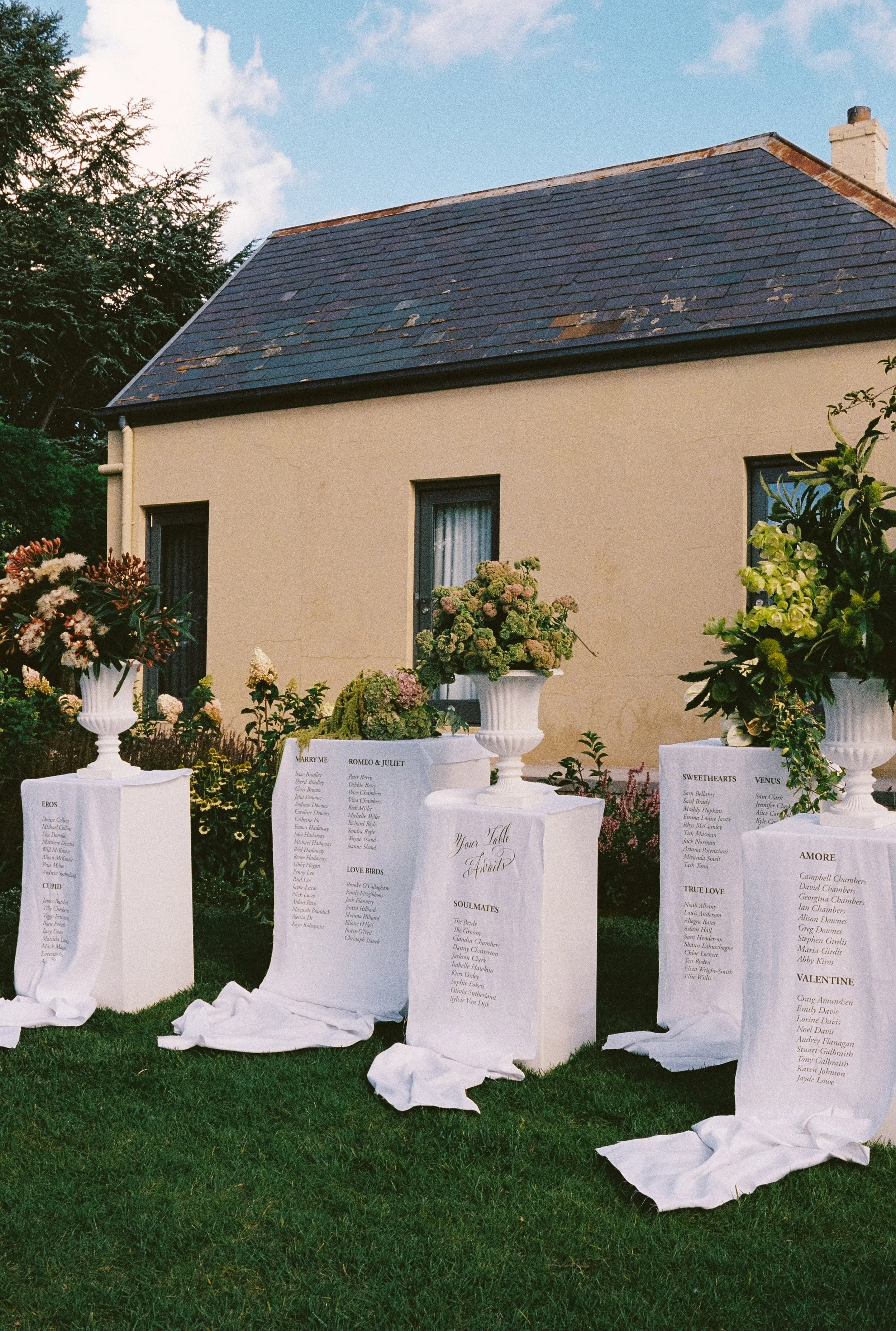 Outdoor wedding or event setup with white fabric drapes displaying lists of names, possibly bridesmaids or guests, set against a house and garden with various plants and flowers.