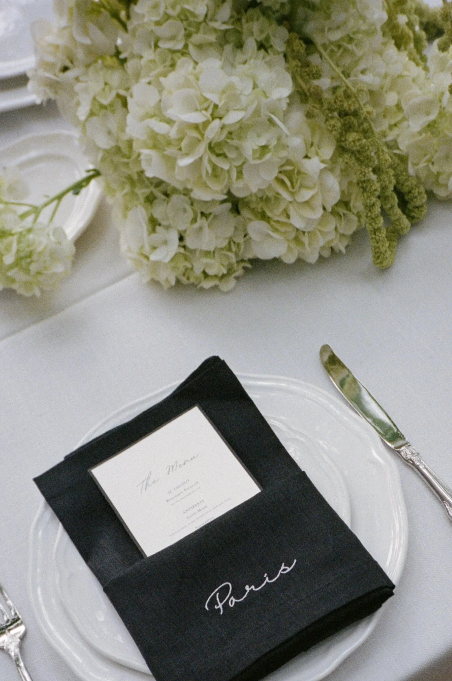 A white hydrangea flower bouquet, a black box with a white label, a silver knife, and a white plate with a glass border on a white tablecloth. The box has the word 'Parties' written on it.