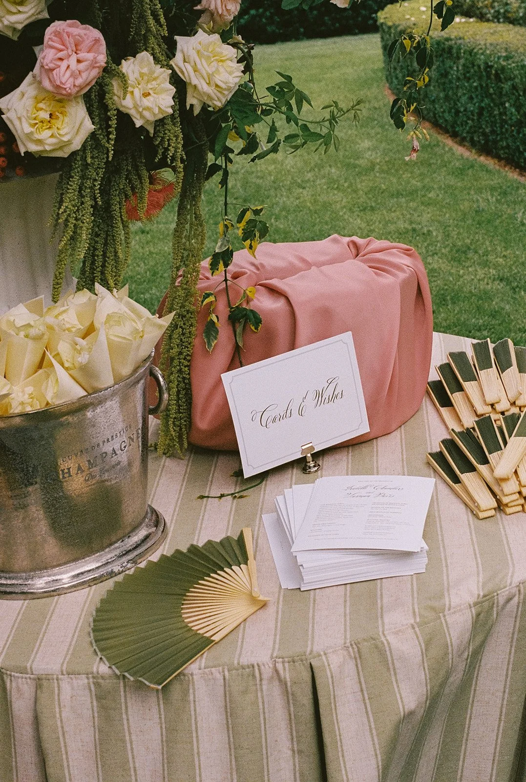 A table at an outdoor wedding reception with a floral arrangement, a pink fabric-covered gift box, and stacks of paper and wooden clothespins for guest books and messages. There are also paper fans on the table.