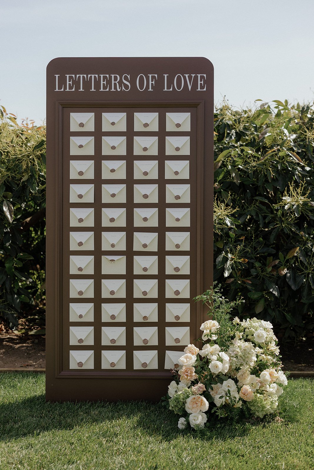 A display board labeled "Letters of Love" with multiple envelopes, and a bouquet of white flowers in front, outdoors with lush green shrubbery in the background.