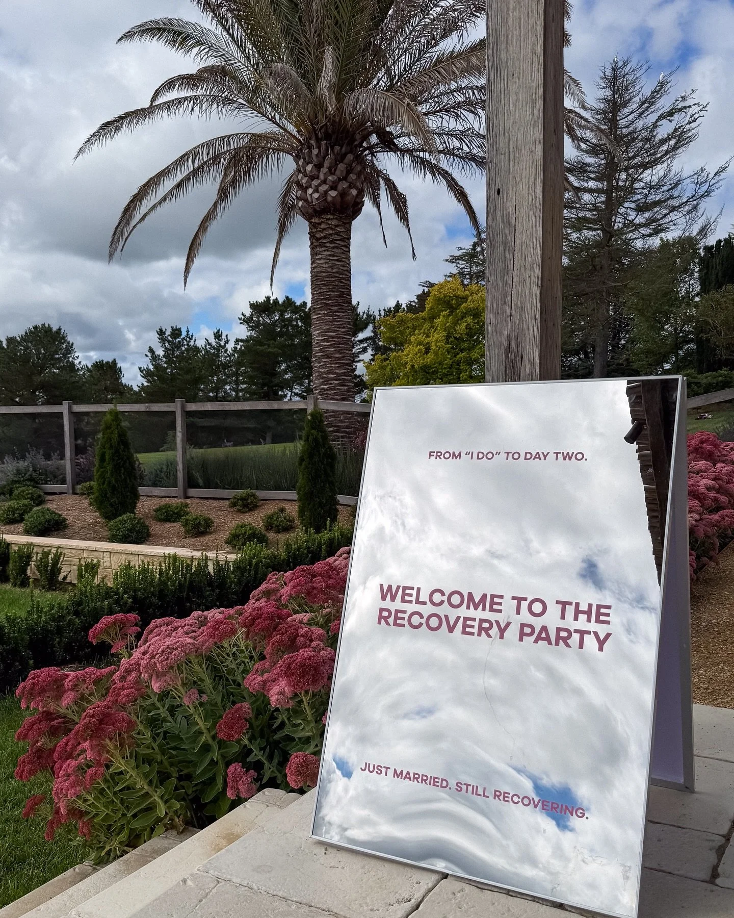 Signboard outdoors at a recovery event with flowers in the foreground and trees, palm tree, and cloudy sky in the background. The sign says 'From I do to day two. Welcome to the recovery party. Just married. Still recovering.'