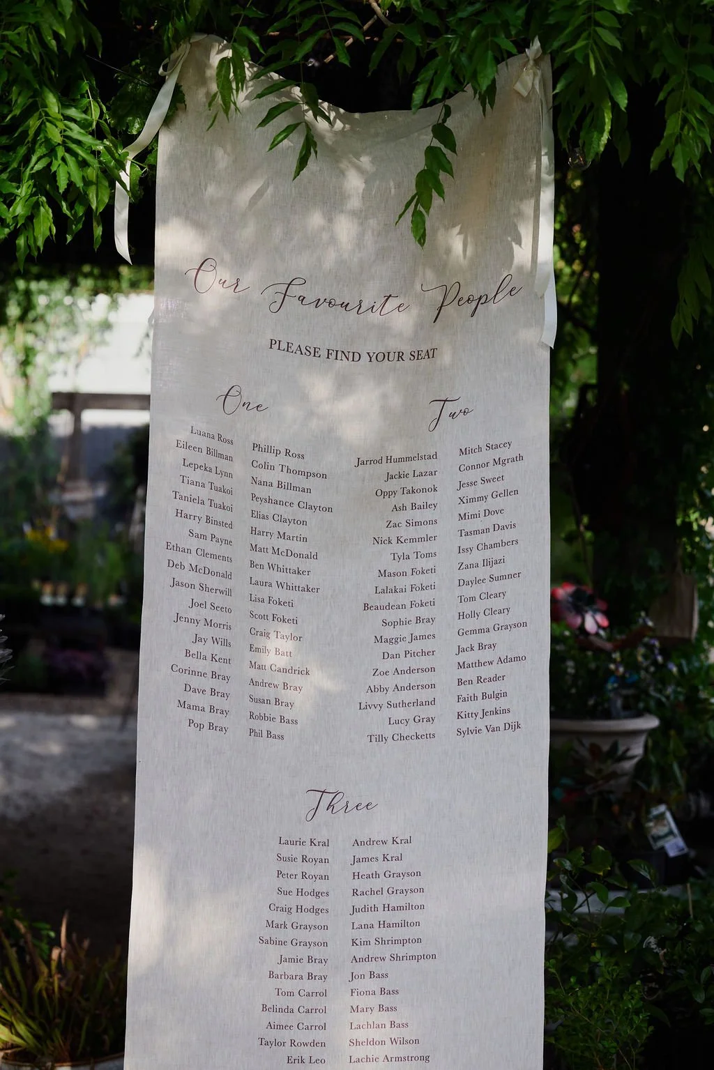 A wedding seating chart listing the names of guests divided into three sections: One, Two, and Three, with the heading 'Our Favourite People.' It is a tall, white fabric sign hung on a tree with white ribbons, outdoors, with greenery and flowers in the background.